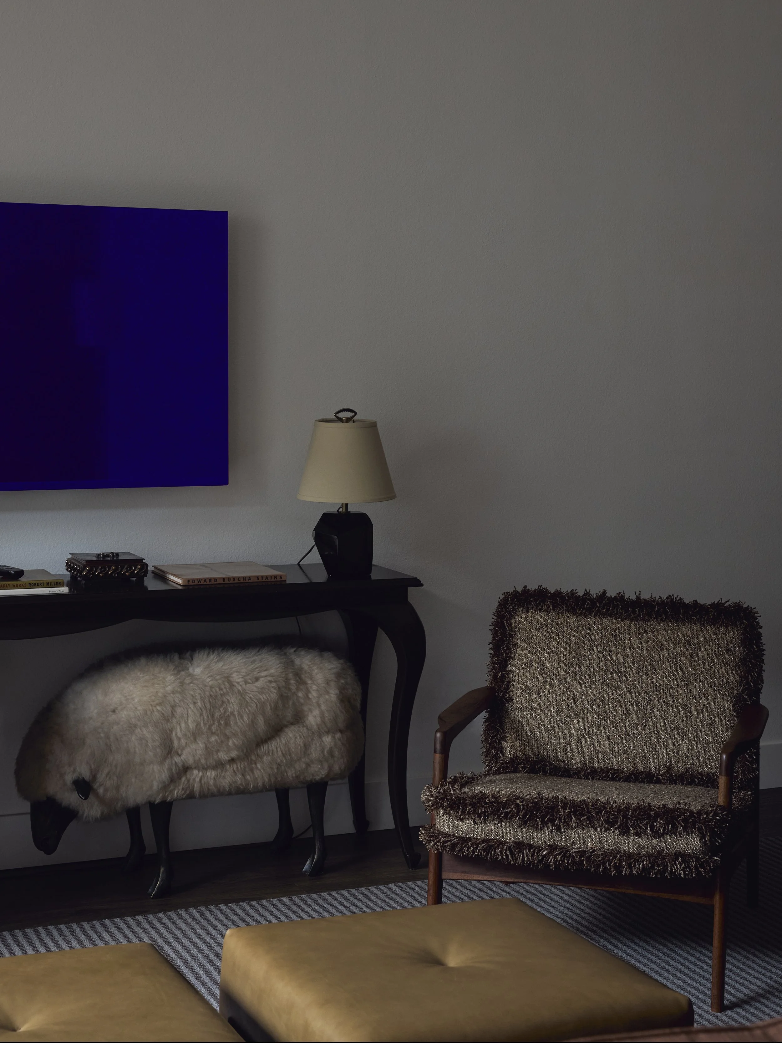 Living room with a black console table, a fur-covered stool with animal legs, a textured armchair, and a mustard-colored ottoman. The wall features a large blue painting, a lamp, and books.