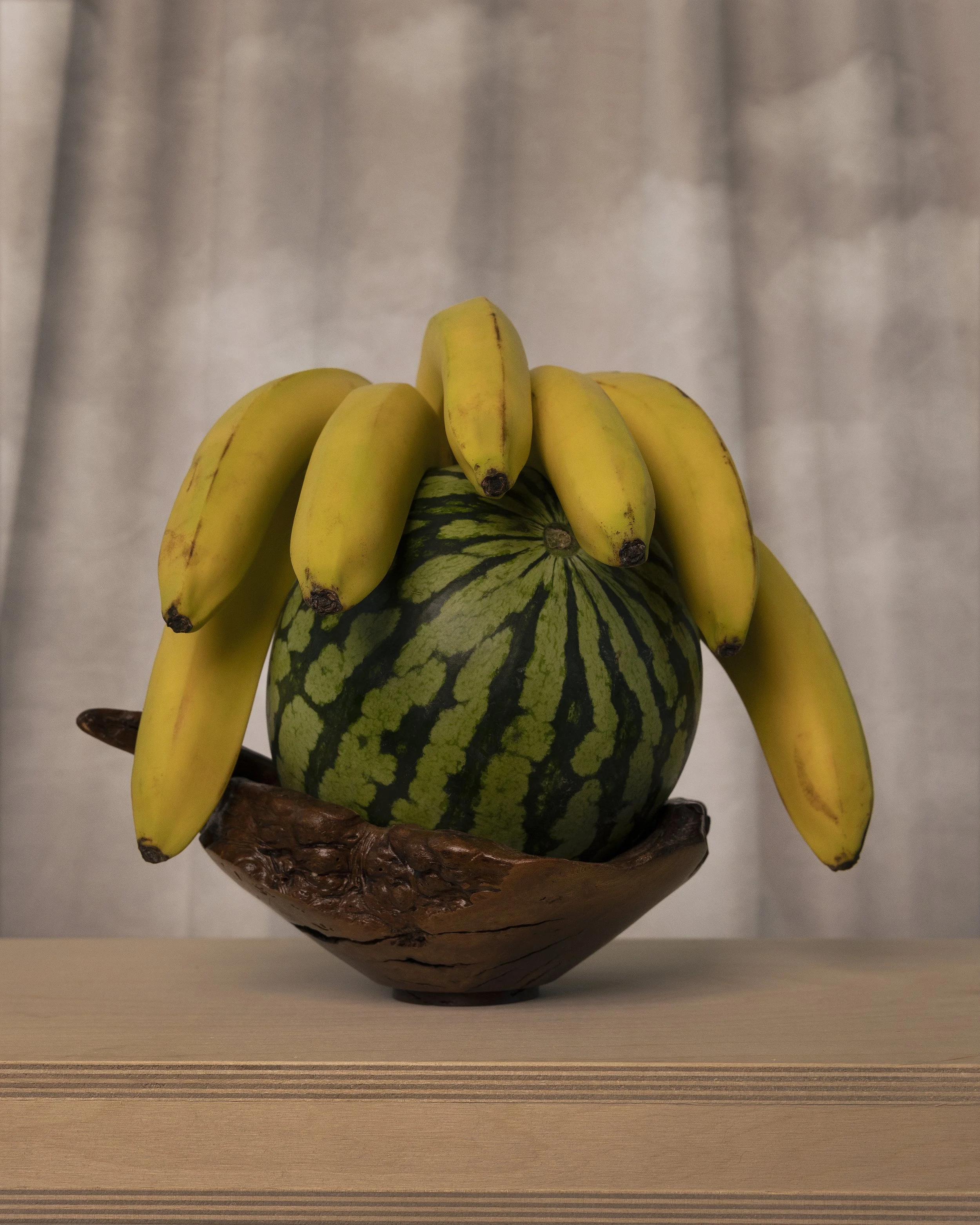 A decorative wooden bowl with a watermelon and a bunch of bananas on top, placed on a wooden surface.