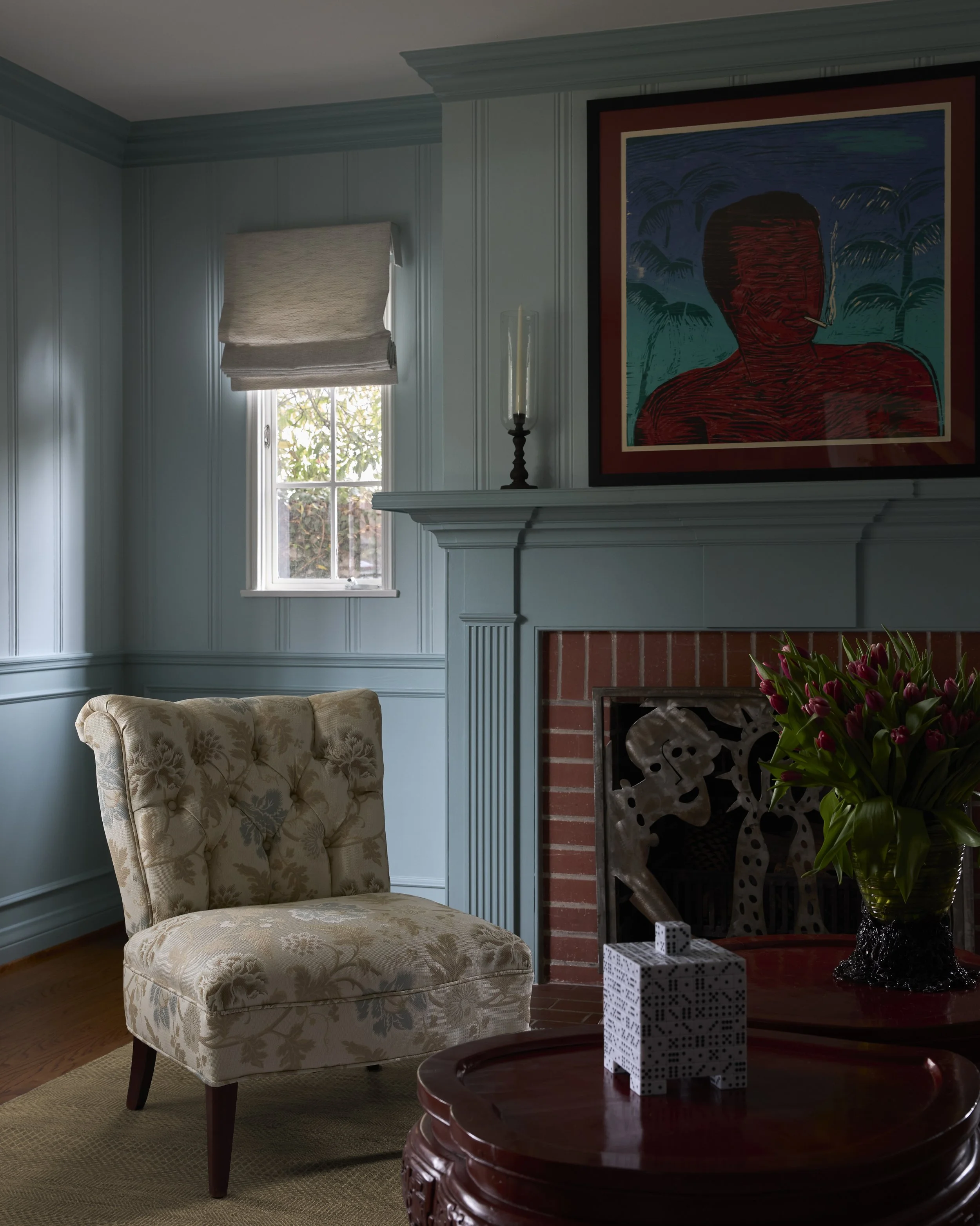 A cozy living room corner with a floral upholstered armchair, a window with a beige roman shade, blue-paneled walls, a decorated white fireplace mantel with a candle in a glass holder, a painting of a faceless person smoking a cigarette, a bouquet of pink flowers on a round wooden table, and a couple of patterned gift boxes on the table.