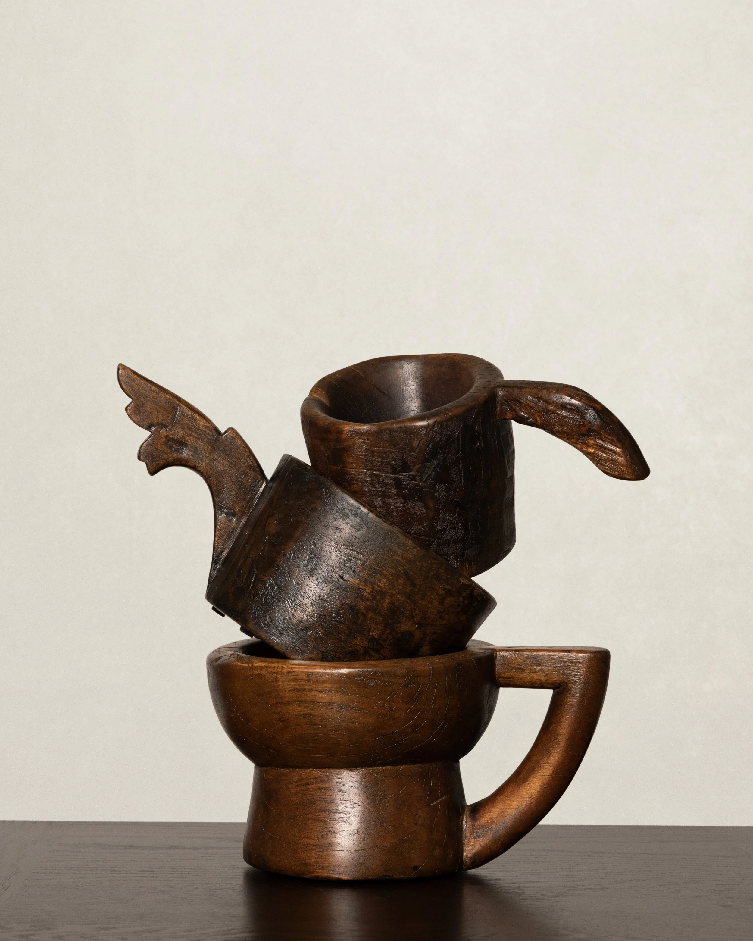 A carved wooden sculpture resembling a traditional mythological creature or figure, with a large, rounded body, a decorated head, and a handle on the right side, placed on a dark wooden surface against a plain, light-colored background.