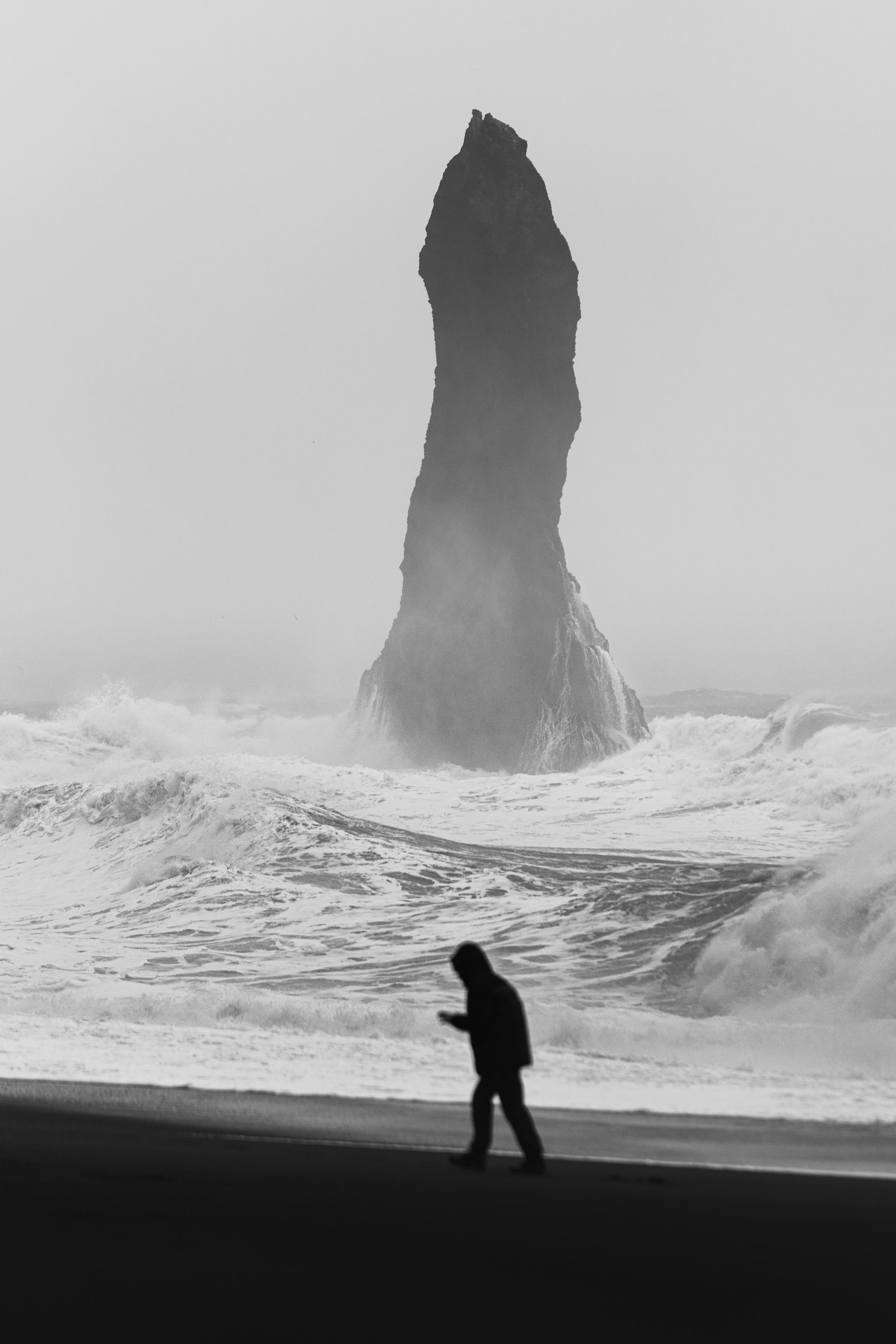 Alone — black and white fine art seascape photograph of a lone figure walking on a black sand beach beneath a towering sea stack and crashing waves, Iceland, by Vasilis Moustakas