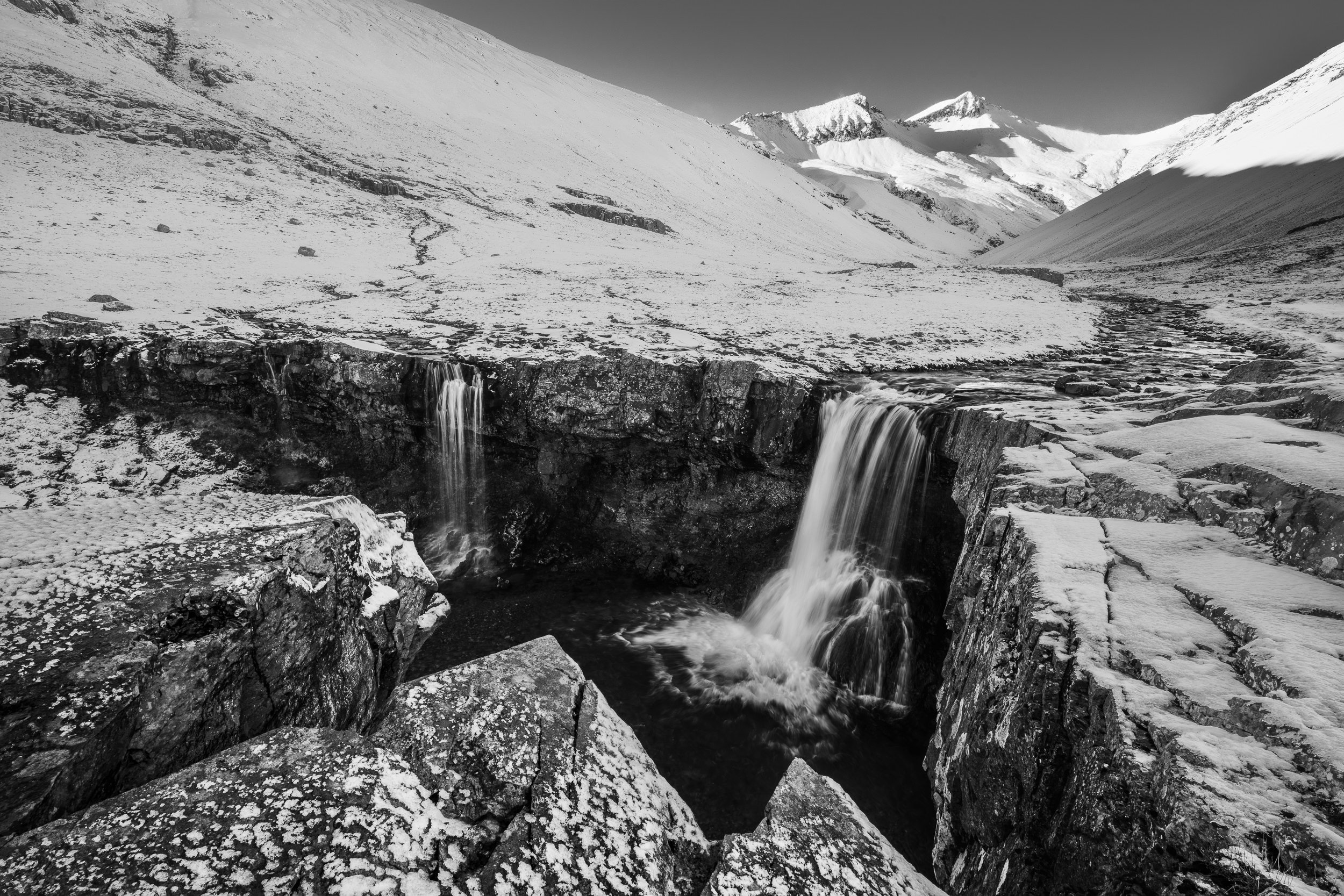 The View — black and white fine art landscape photograph of a waterfall cascading over rocky ledges into a dark pool surrounded by snow-covered mountains, Iceland, by Vasilis Moustakas
