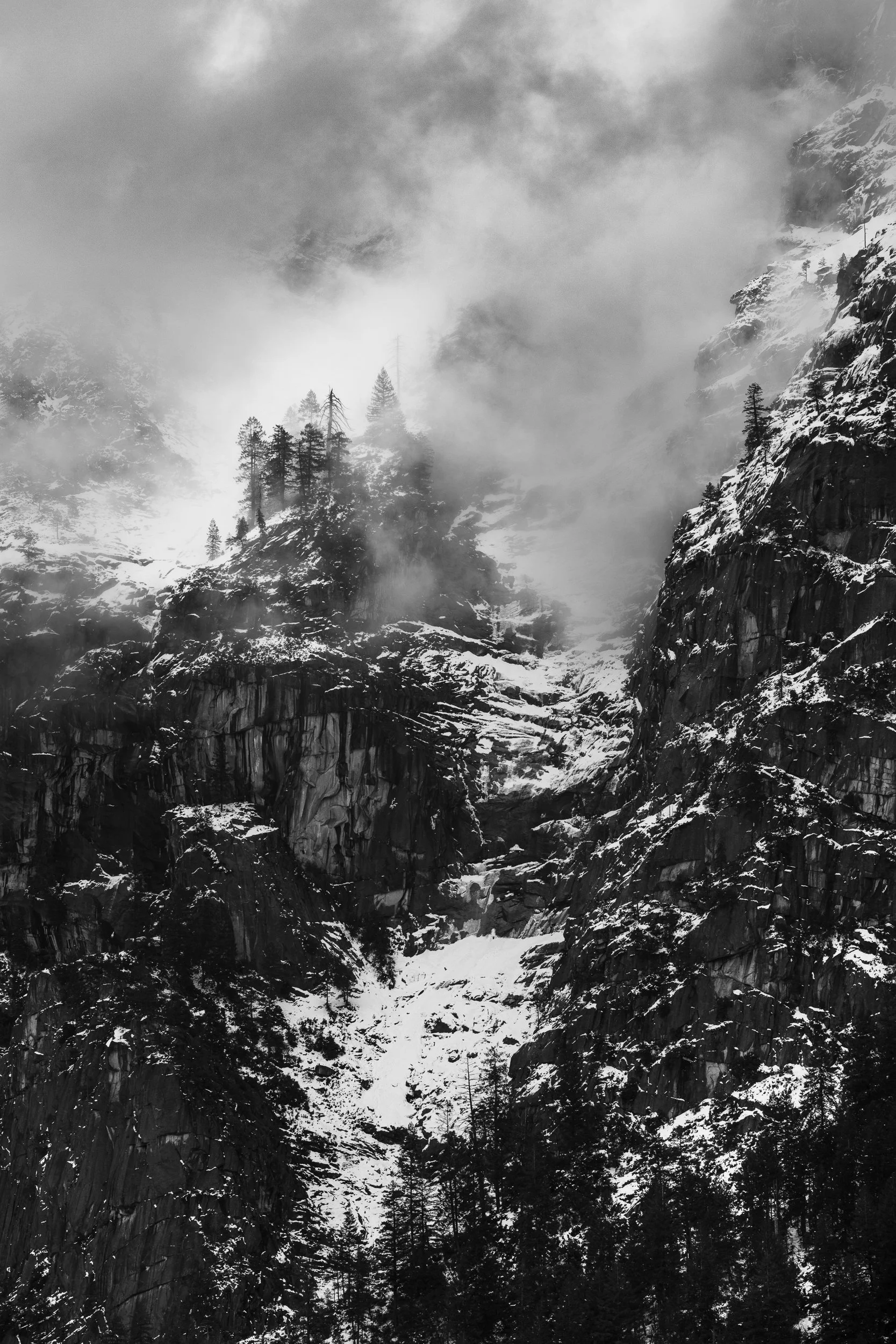 Dramatic black and white photograph of granite cliffs in Yosemite Valley with snow, mist, and pine trees during early spring weather.