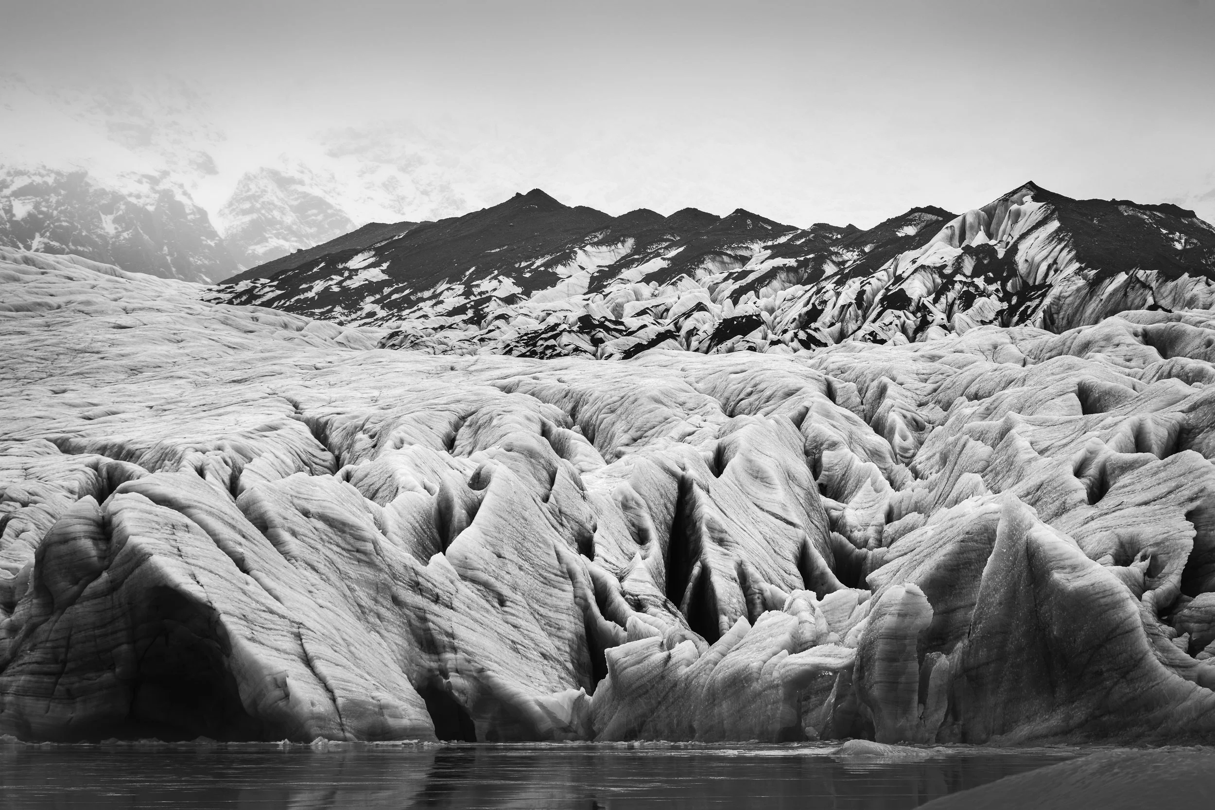 Glacier's Song — black and white fine art landscape photograph of deeply crevassed glacier ice with snow-covered mountains beyond, Iceland, by Vasilis Moustakas