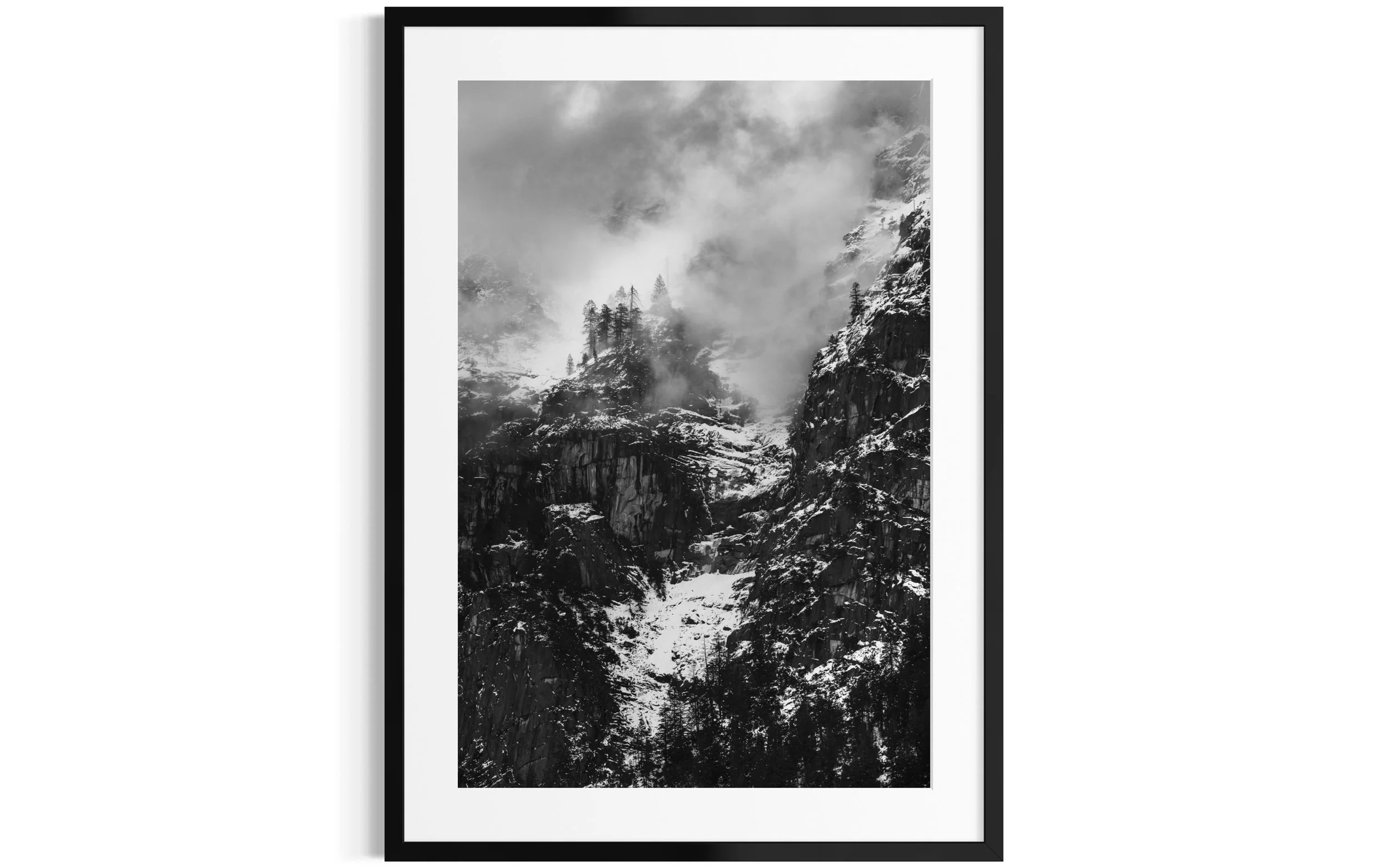 Black and white photograph of a mountain landscape with rocky cliffs, snow, trees, and clouds.