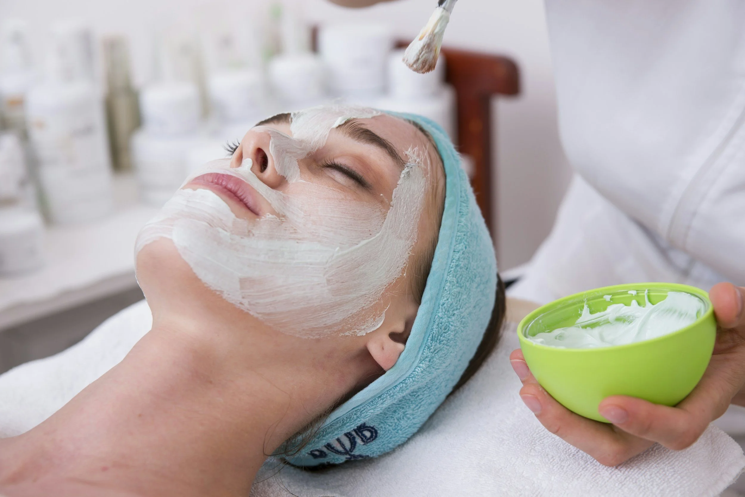 Woman getting a facial treatment in a spa, with a creamy mask on her face, lying on a massage table, and a skincare professional holding a bowl of facial mask.
