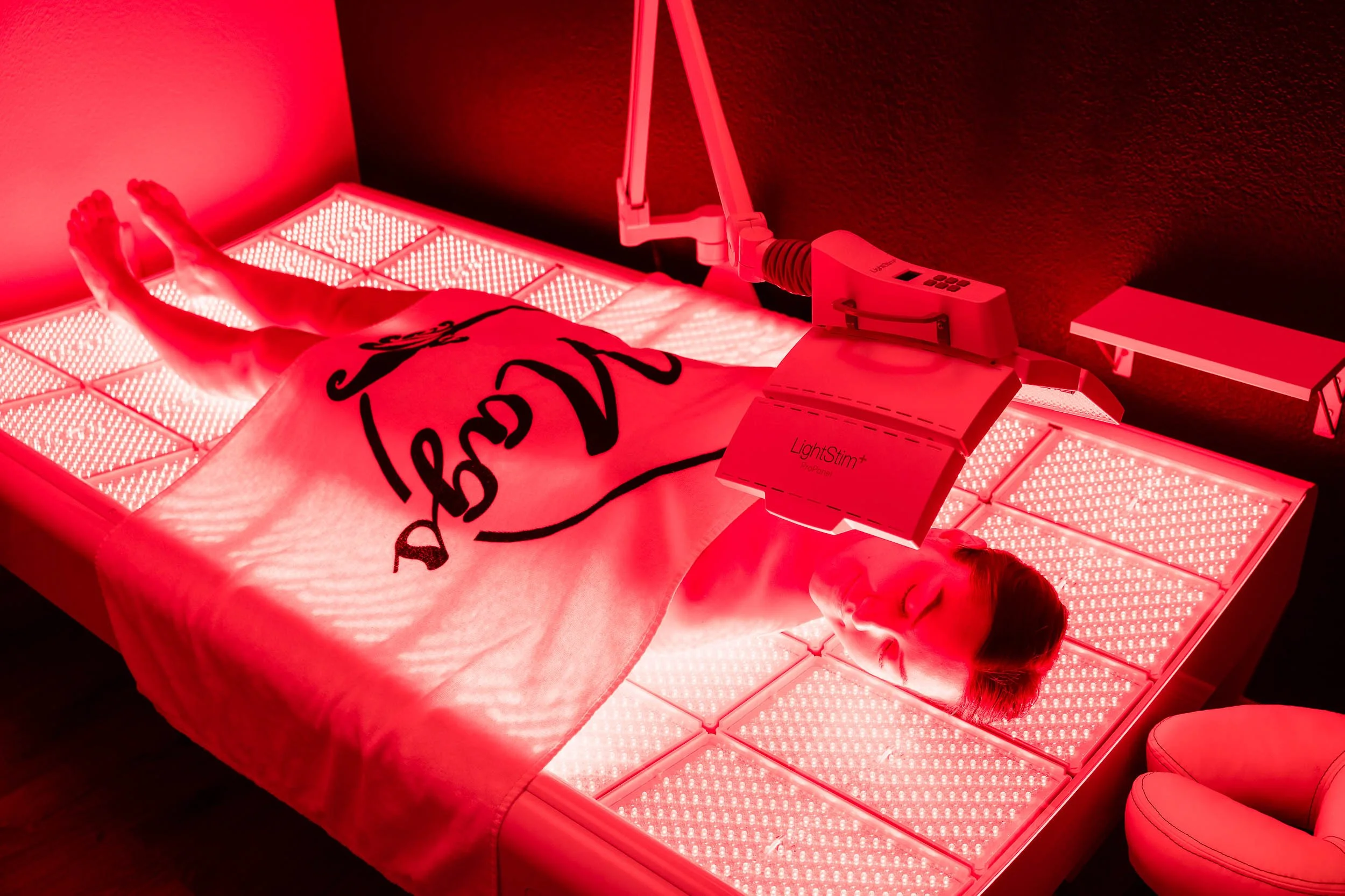 Caucasian female laying flat on a LightStim Red Light therapy bed covered in a wash of red light. She is covered with a Mags Cathey Spa white towel.