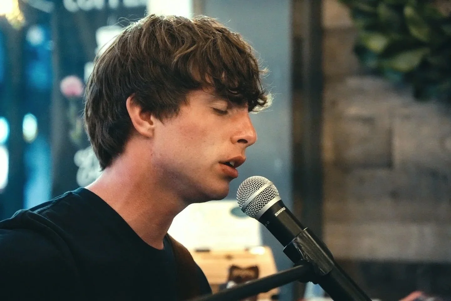 A young man with curly brown hair singing into a microphone with his eyes closed.