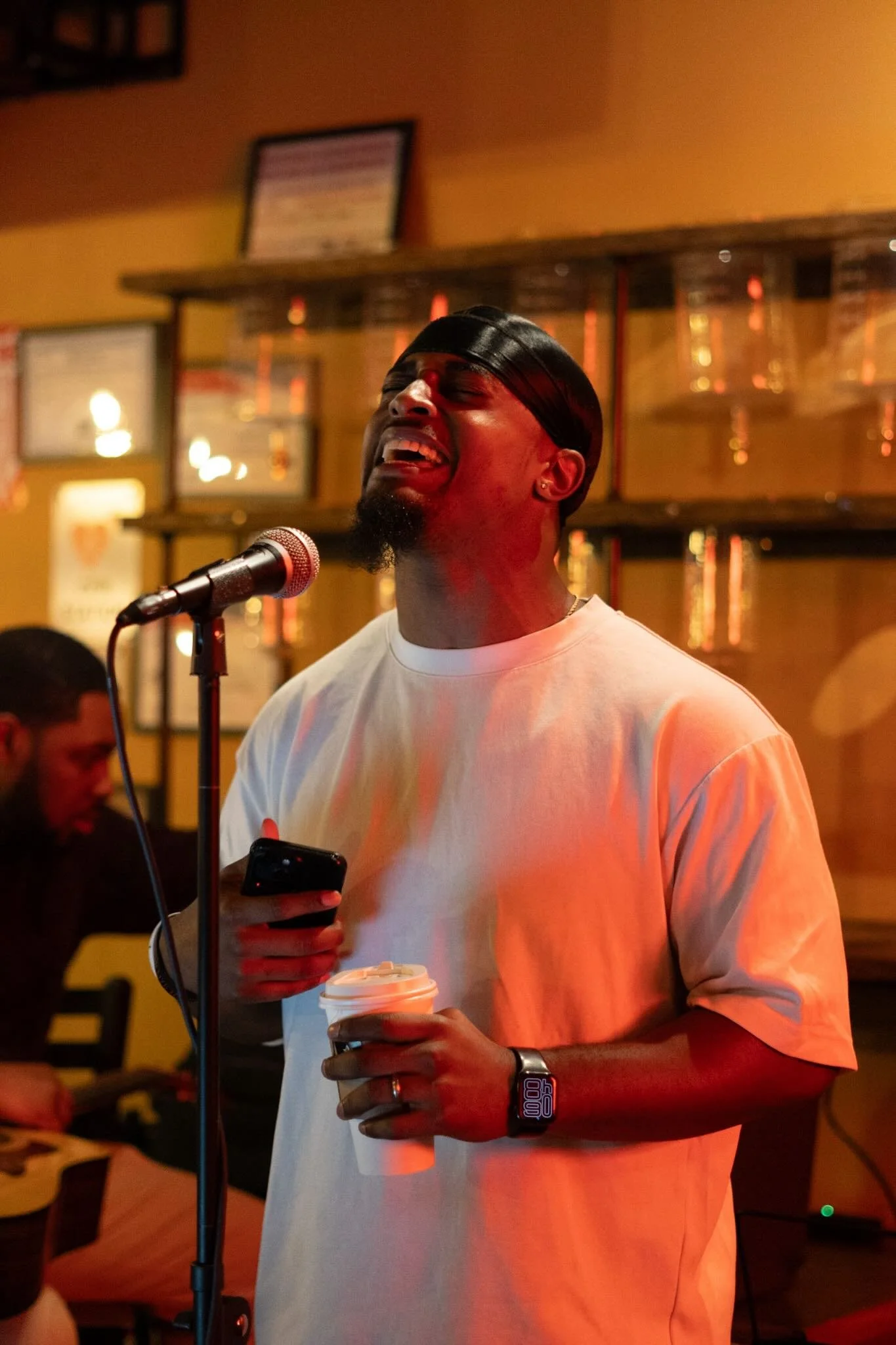 A man singing passionately with eyes closed, holding a coffee cup in one hand and a phone in the other, in a dimly lit bar or cafe.