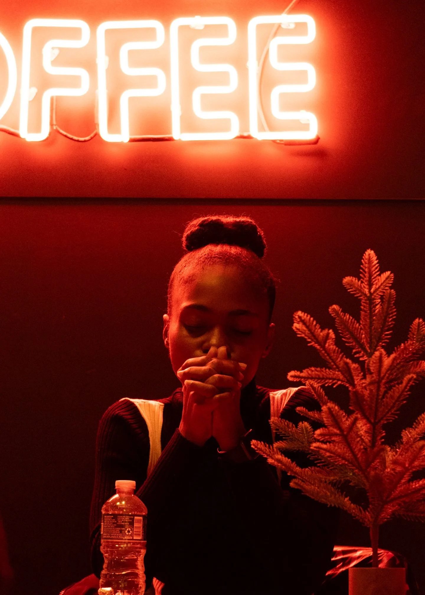 A woman with eyes closed and hands clasped in front of her face sits in front of a neon sign that says 'PEACE'. She is inside a room with dark walls, a small tree in a pot, and a water bottle on the table.