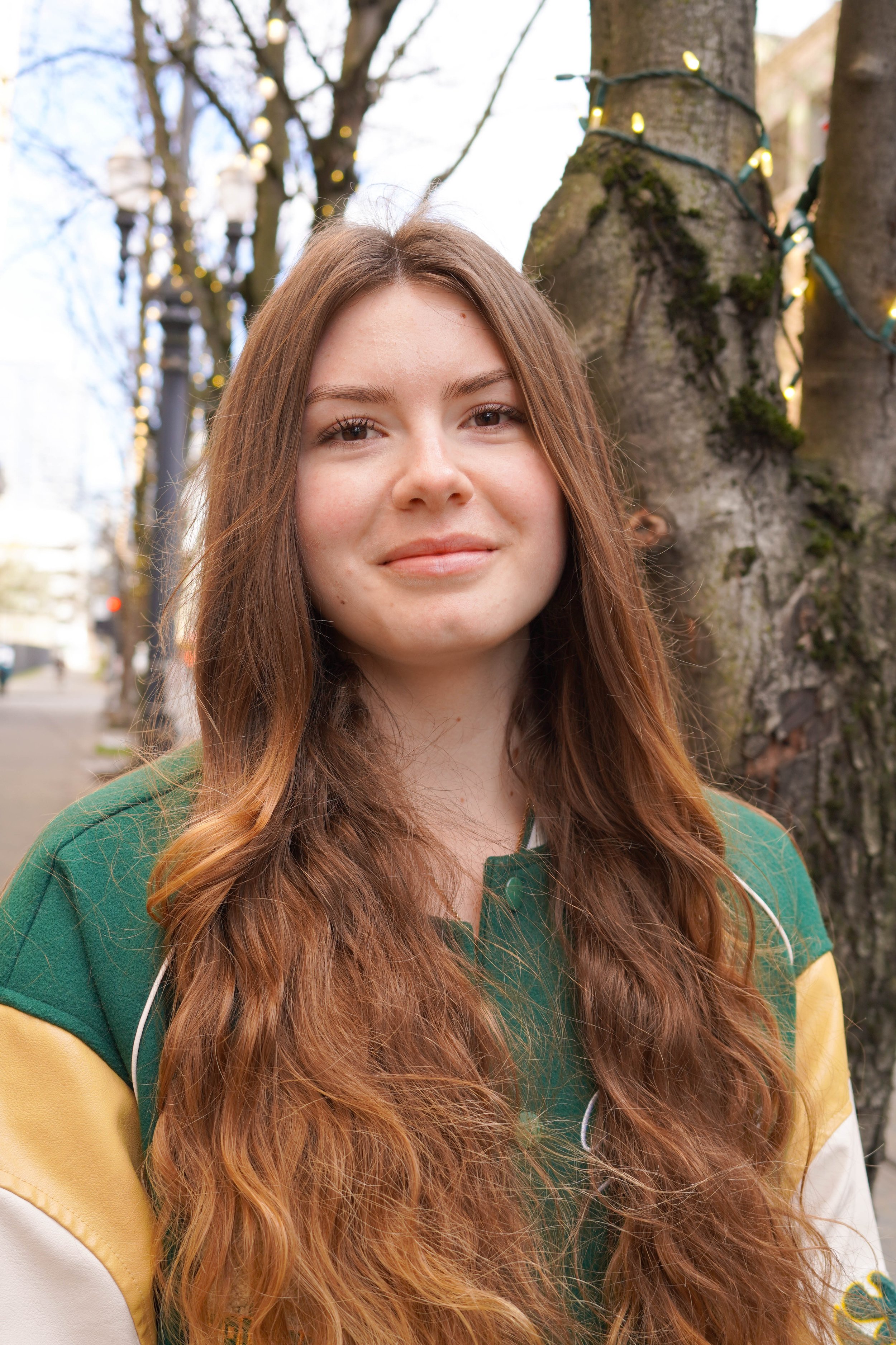 A young woman with long wavy hair and a slight smile posing outdoors with trees in the background.