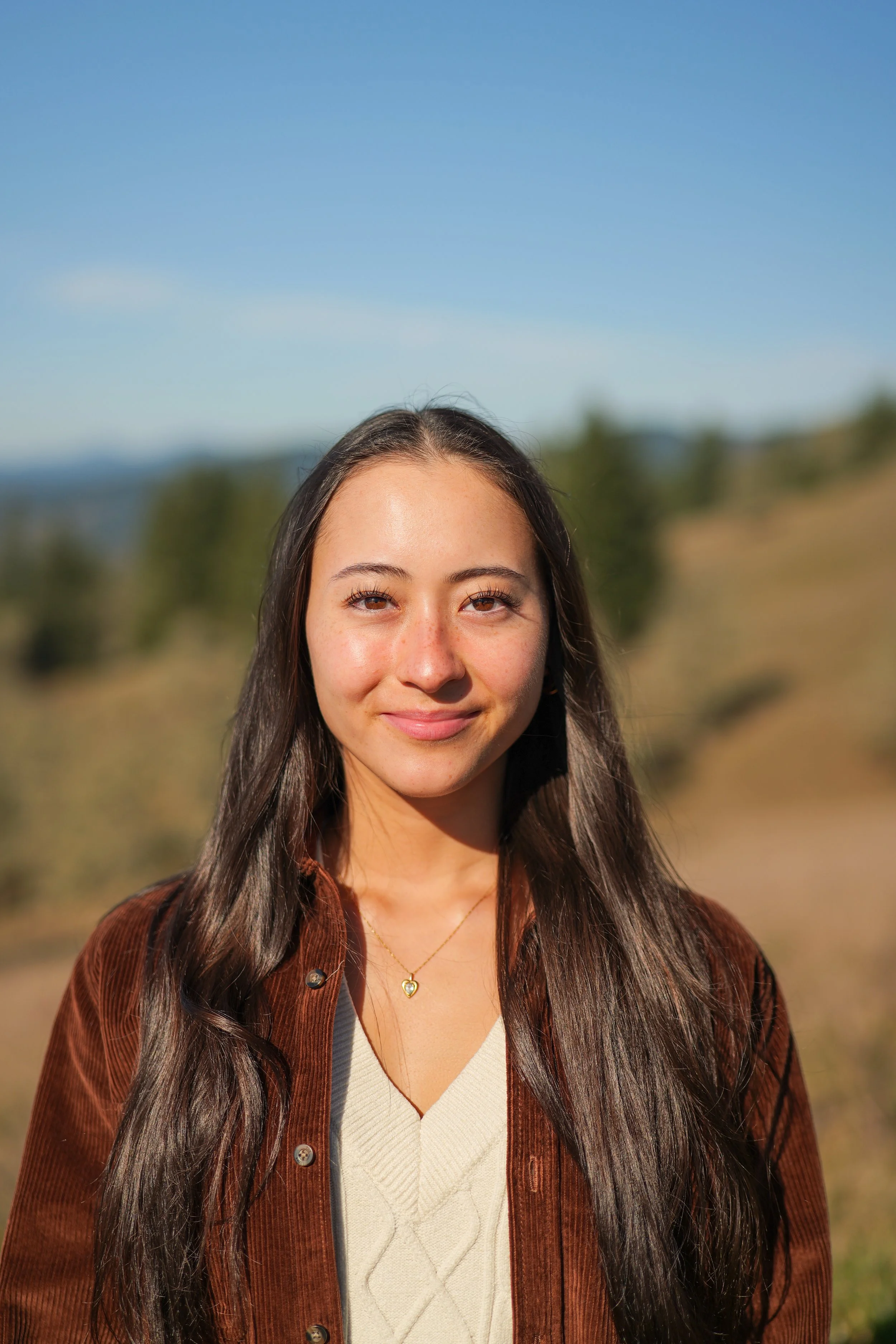 Black and white portrait of a young woman with long dark hair outdoors, wearing a light-colored top, a necklace with a heart-shaped pendant, and a dark jacket.