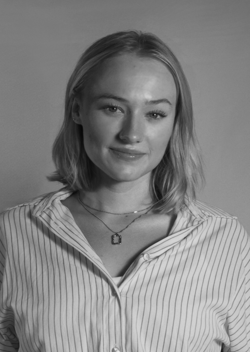 A young woman with shoulder-length blonde hair, wearing a striped shirt and layered necklaces, smiling at the camera in a black and white photo.