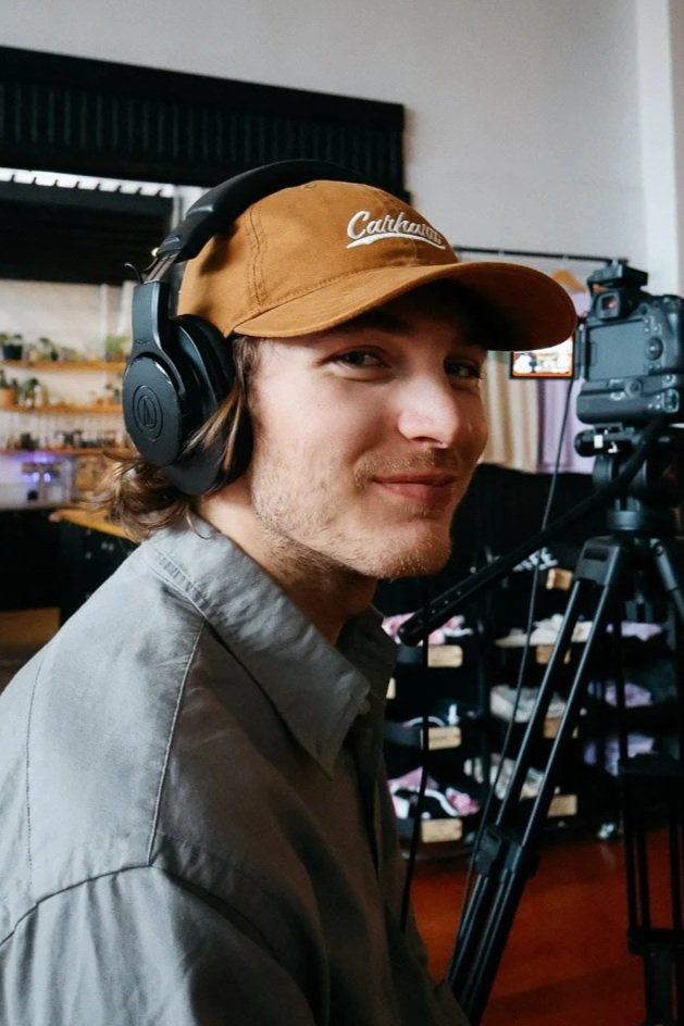 A young man with curly hair smiling in front of a hedge, wearing a button-up shirt with a Polo logo and a necklace.