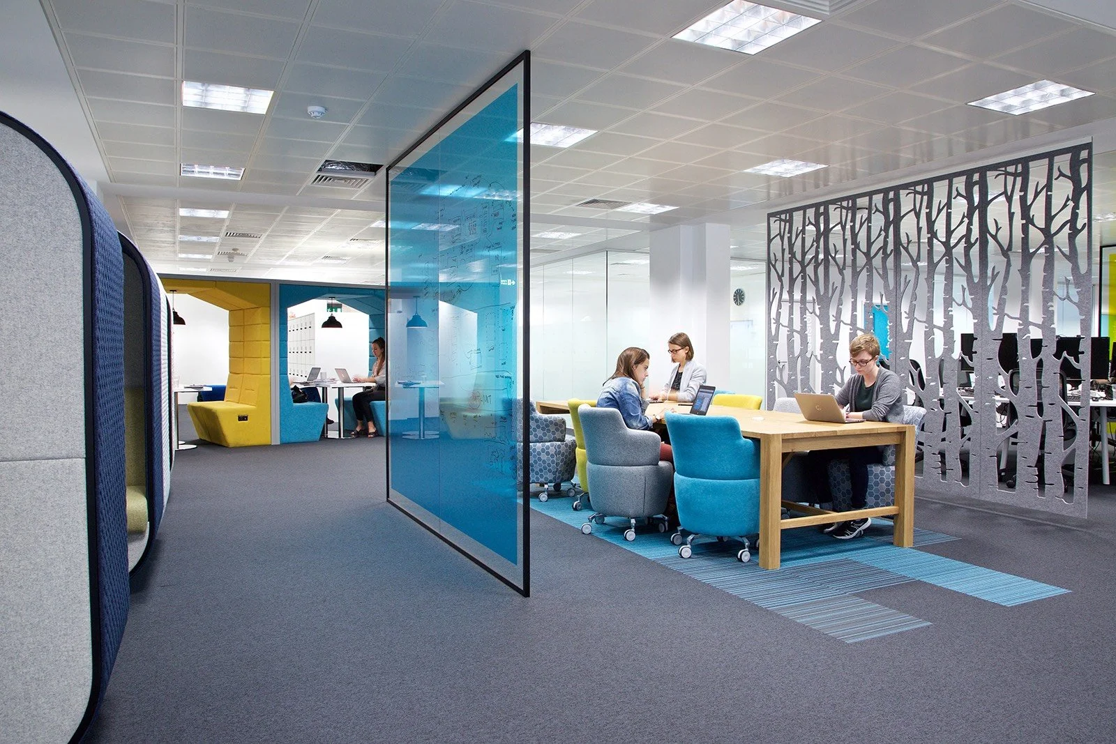 Modern office space with three women working at a wooden table with laptops, separated by decorative panels, with additional workspaces and seating in the background.