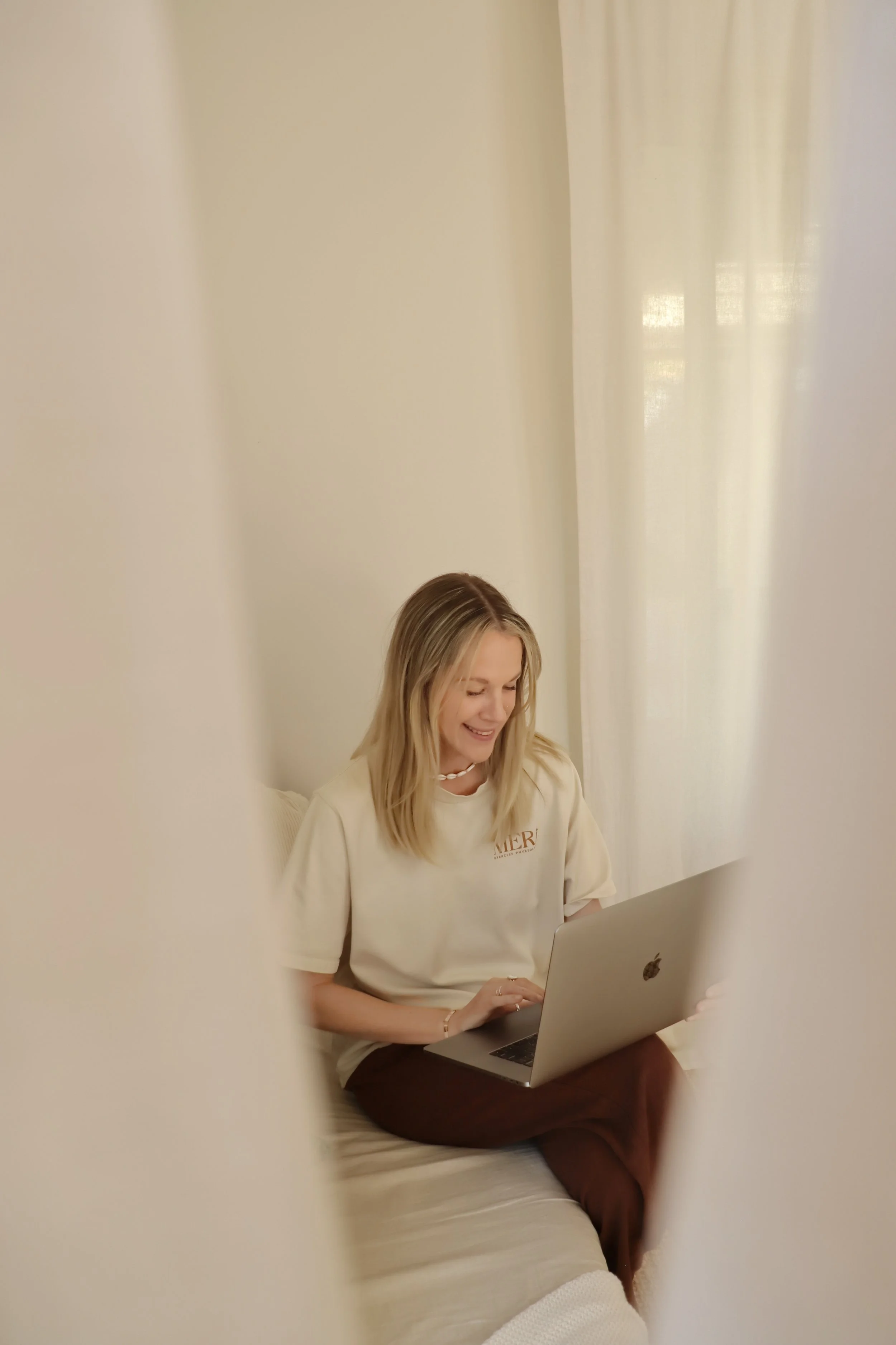 A laptop on a wooden table displays a woman in a yoga pose with hands together in front of her chest, indoors with a brick fireplace and plants in the background.