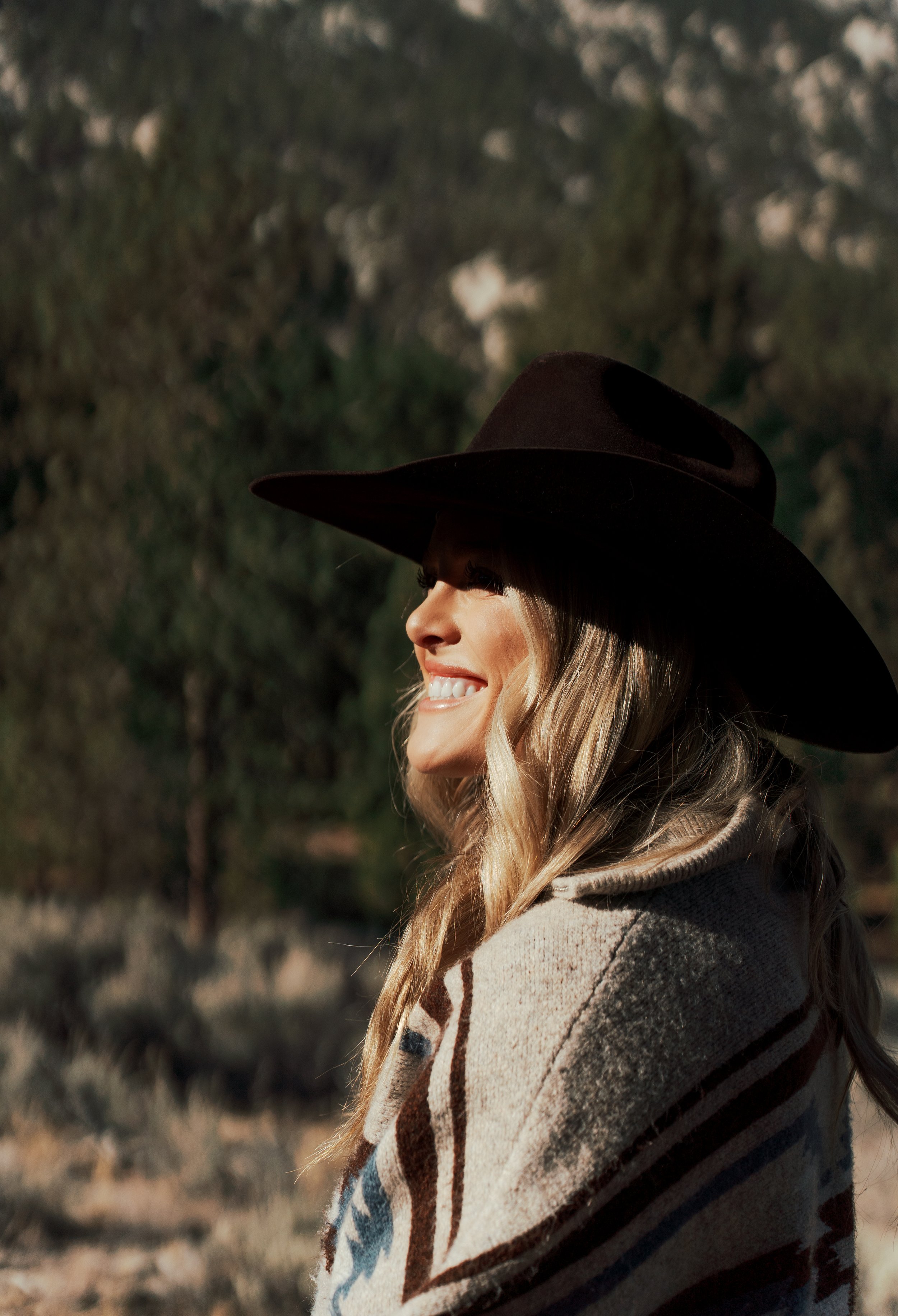 Woman wearing a cowboy hat and a patterned shawl, smiling in a sunny outdoor setting with trees in the background.