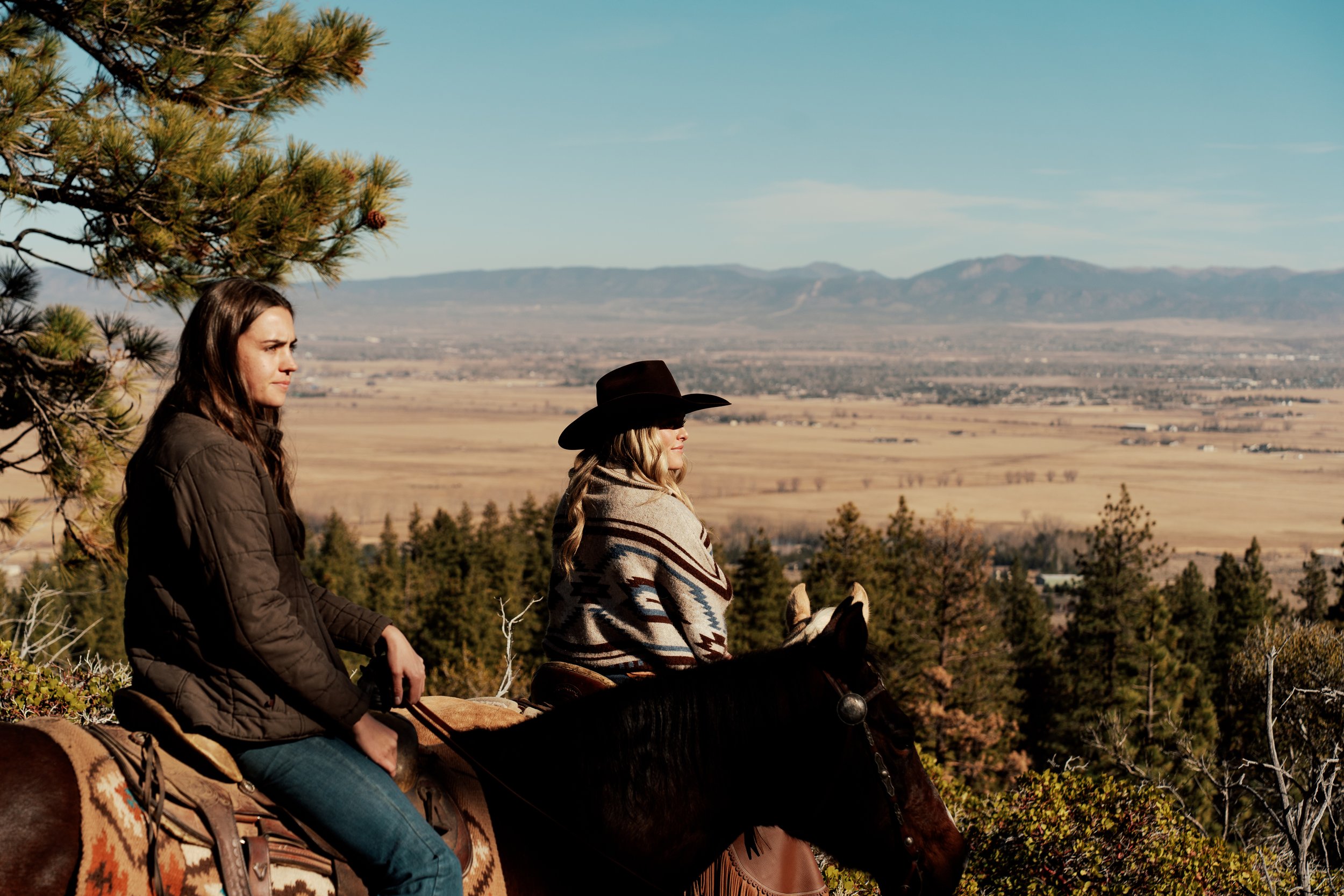 Two people horseback riding on a scenic view overlooking a valley and mountains, with trees in the foreground and a clear blue sky.