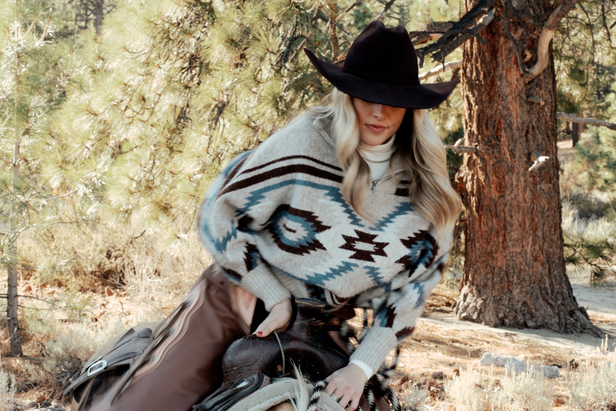 Person in cowboy hat with Western-style sweater and leather pants preparing to saddle a horse in a forest setting.
