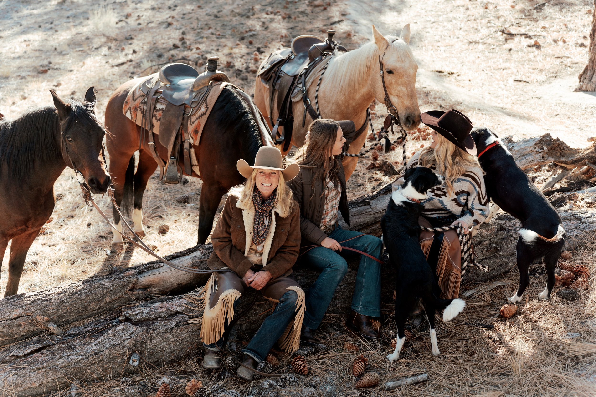 Two people dressed in Western attire sitting on a fallen log with two Border Collie dogs and three saddled horses in a wooded area.