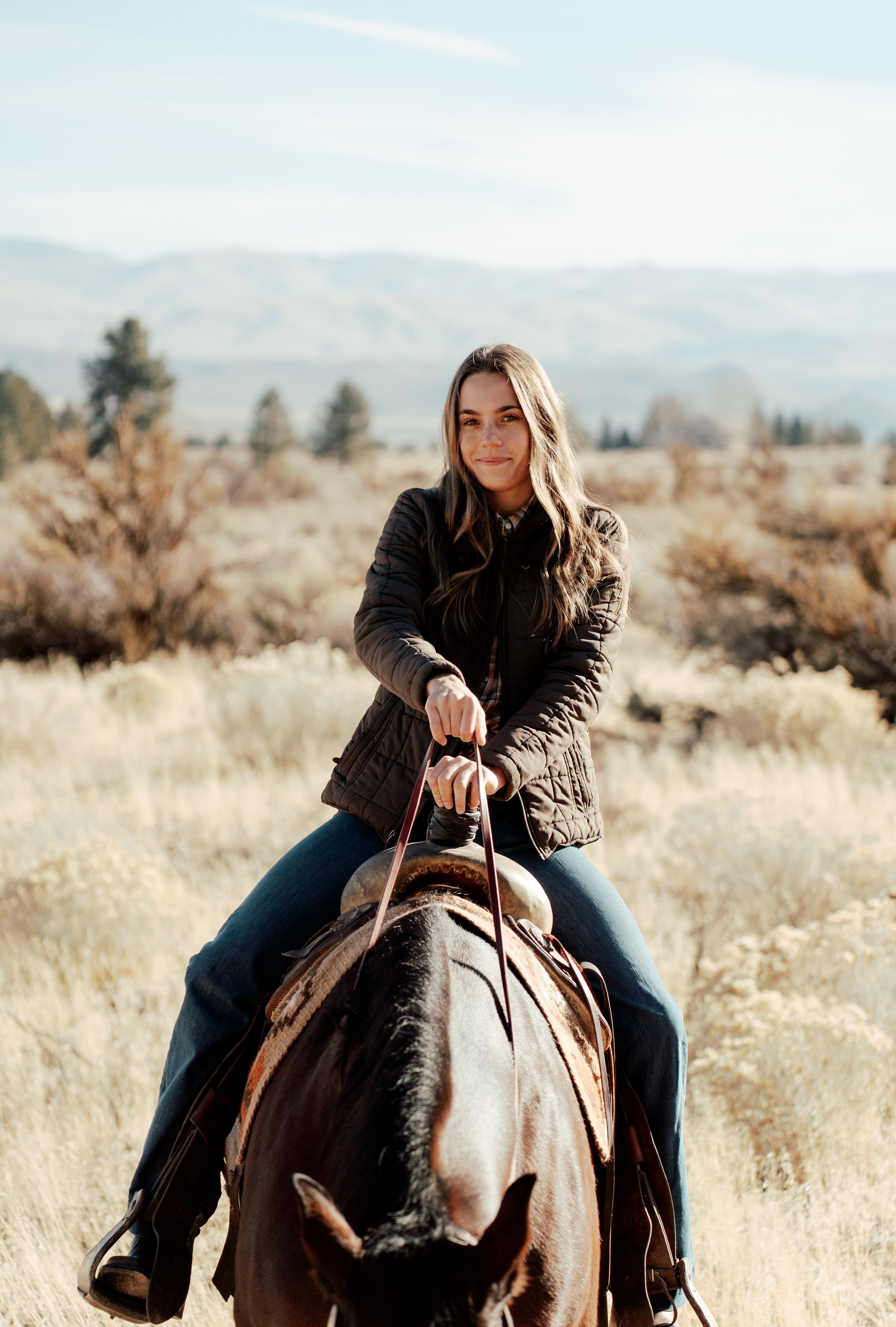 Woman riding a horse in a grassy landscape with distant mountains.