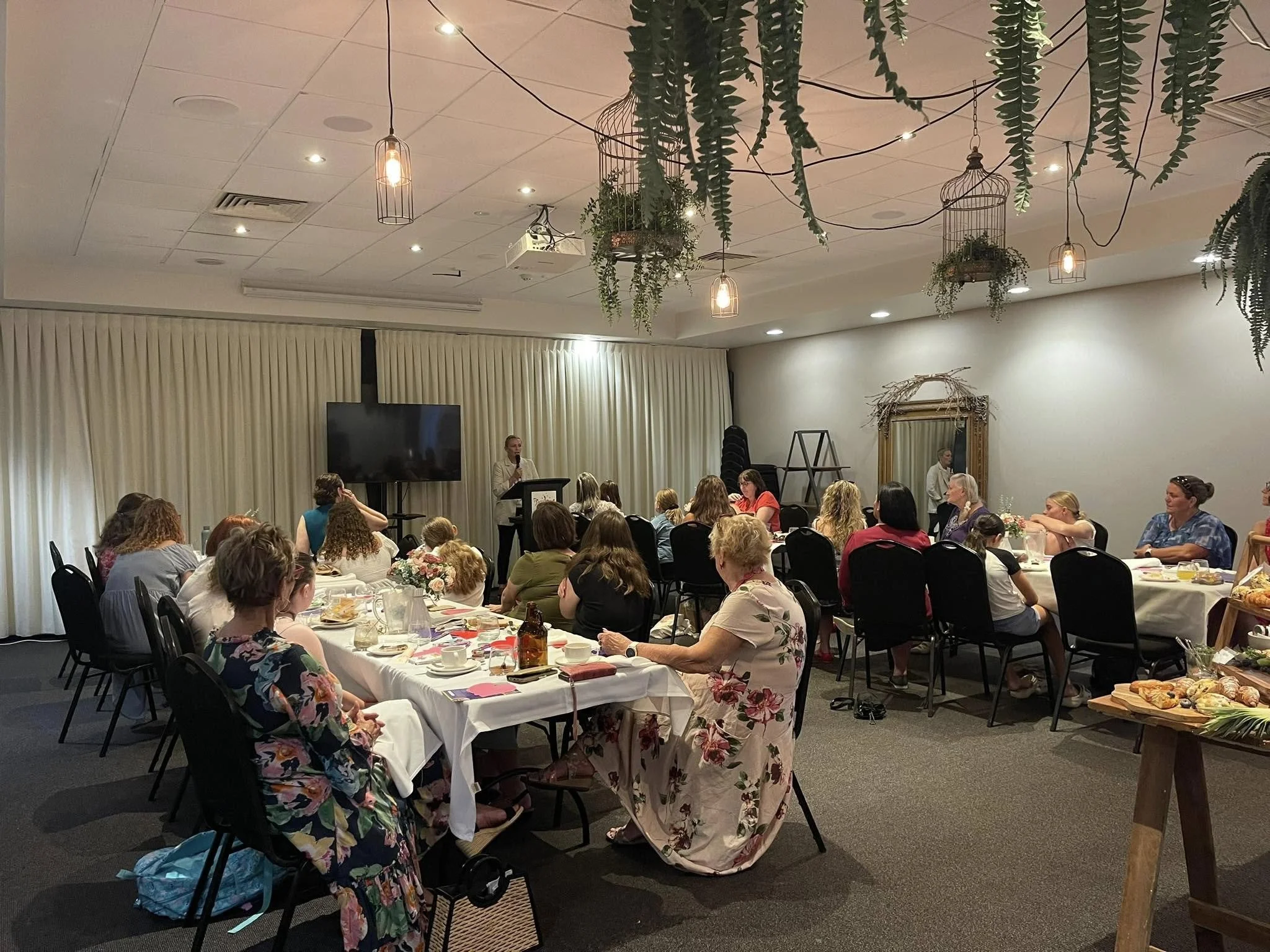 A woman speaking at a podium in front of a room filled with seated women attending a gathering or event. The room is decorated with hanging plants and a large flat-screen TV.
