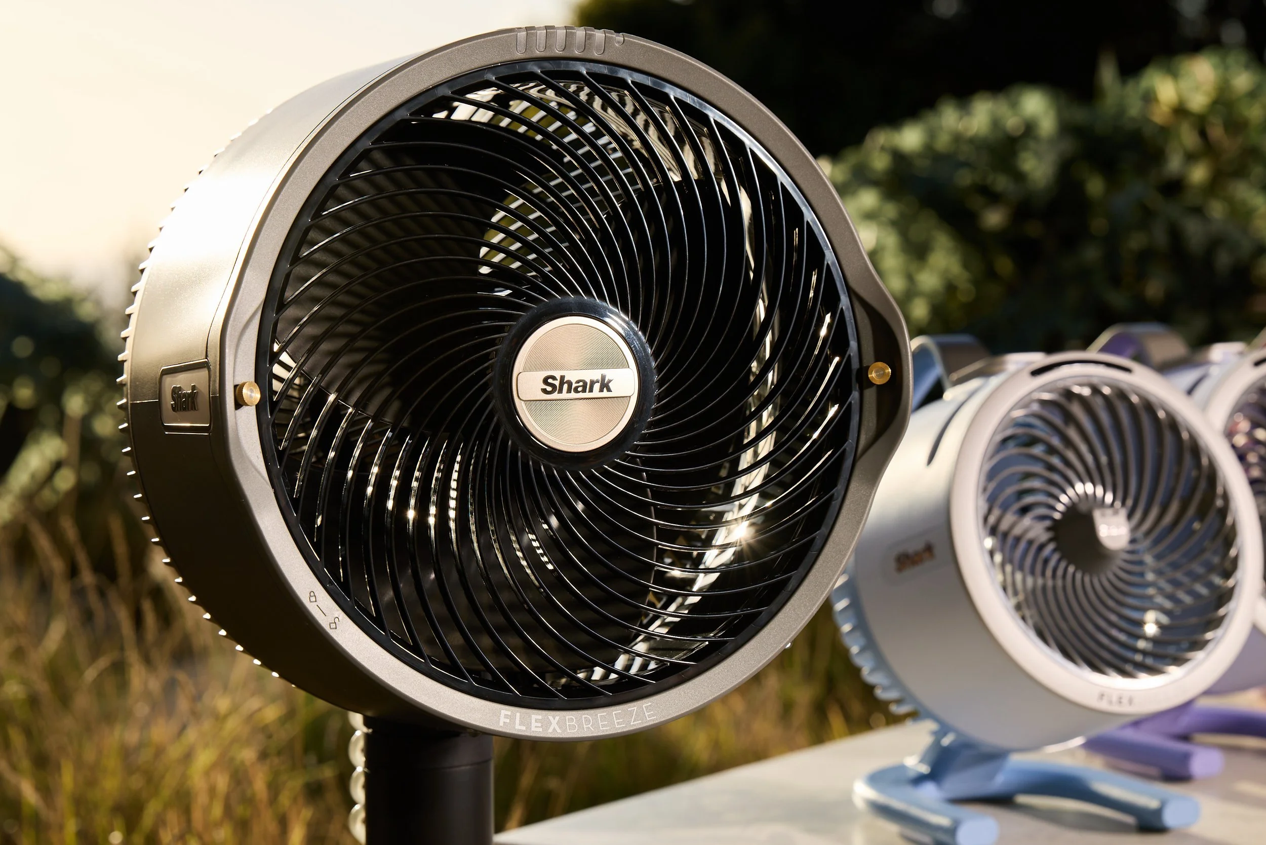Close-up of a black Shark Flex Breeze fan outdoors, alongside other fans in the background, with sunlight and greenery.
