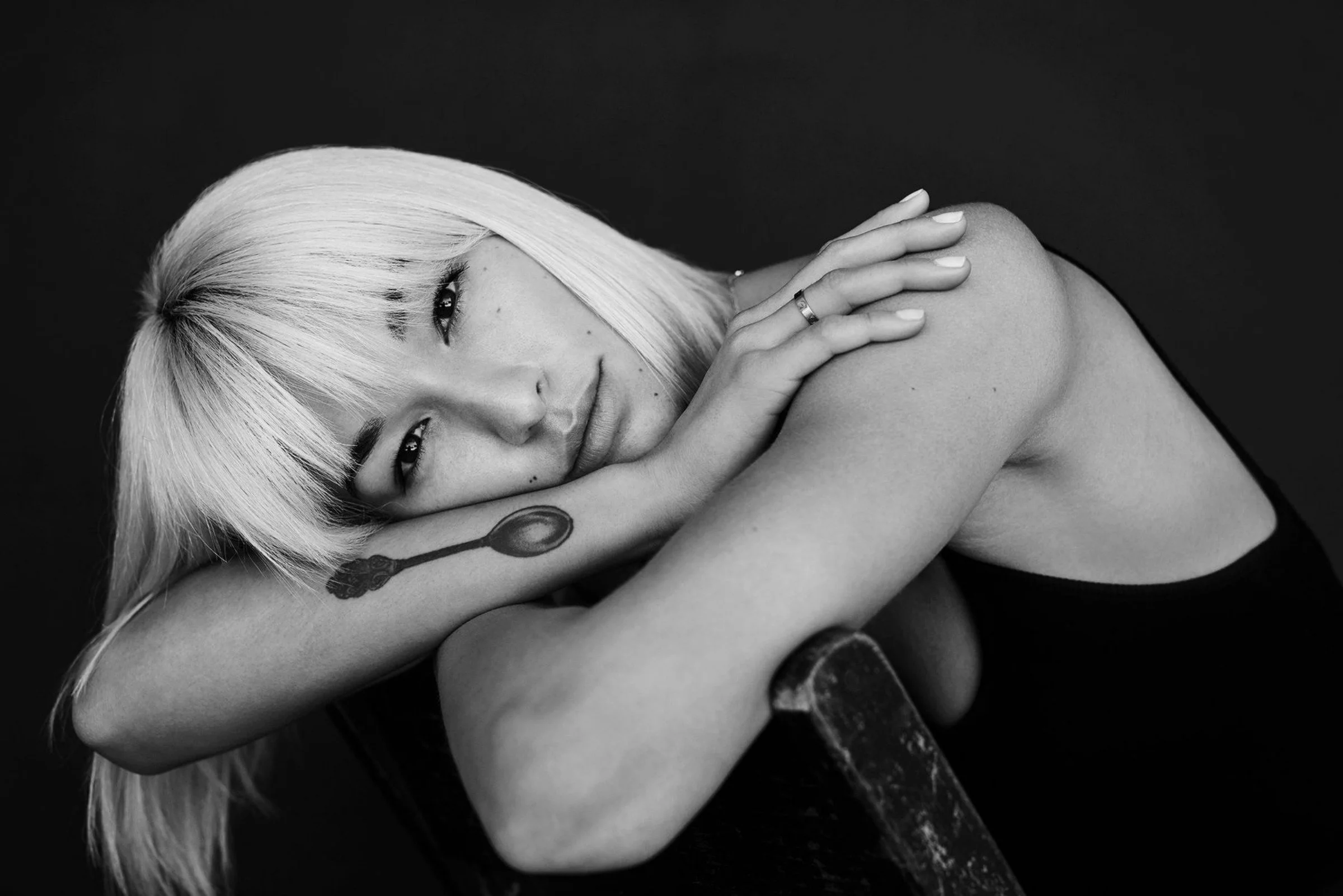 Black-and-white portrait of a blonde woman resting her head on folded arms, with a spoon tattoo visible on her forearm.