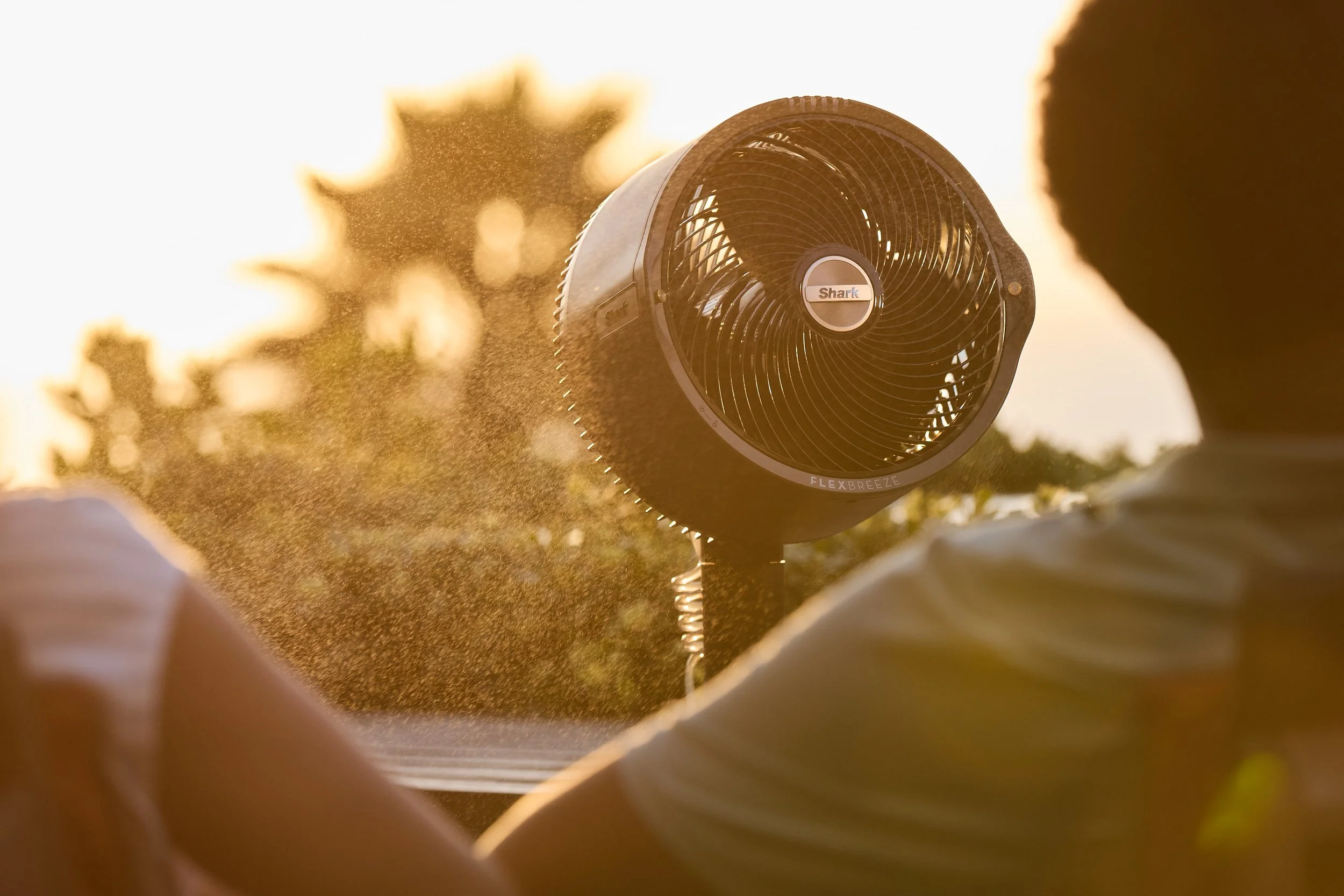 Two people sitting near an outdoor fan emitting mist with sunlight in the background.