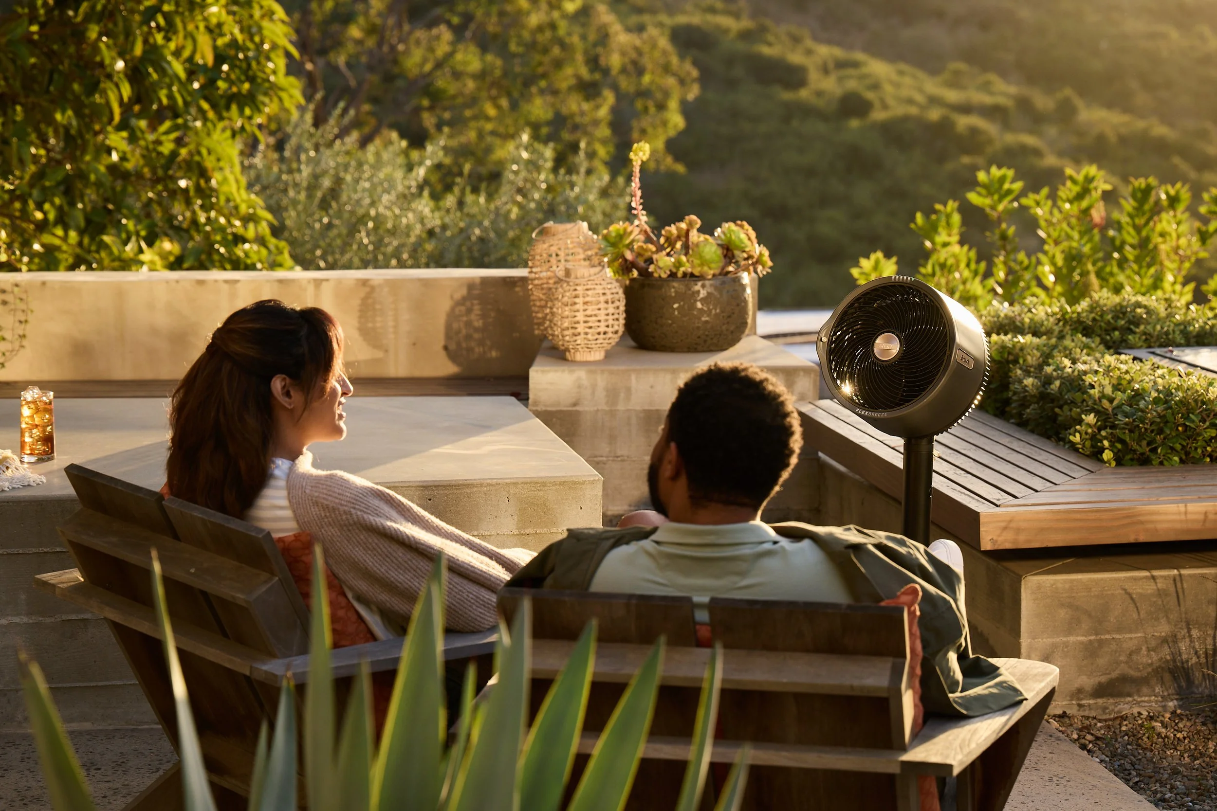 Two people sitting on an outdoor patio with a fan, potted plants, and a glass of iced tea, enjoying a view of greenery in the background.