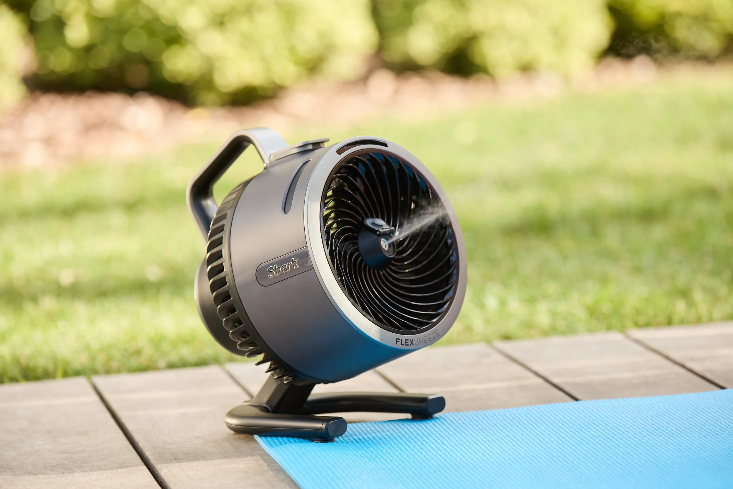 Portable black fan on a wooden deck with a blue yoga mat and green grass in the background.