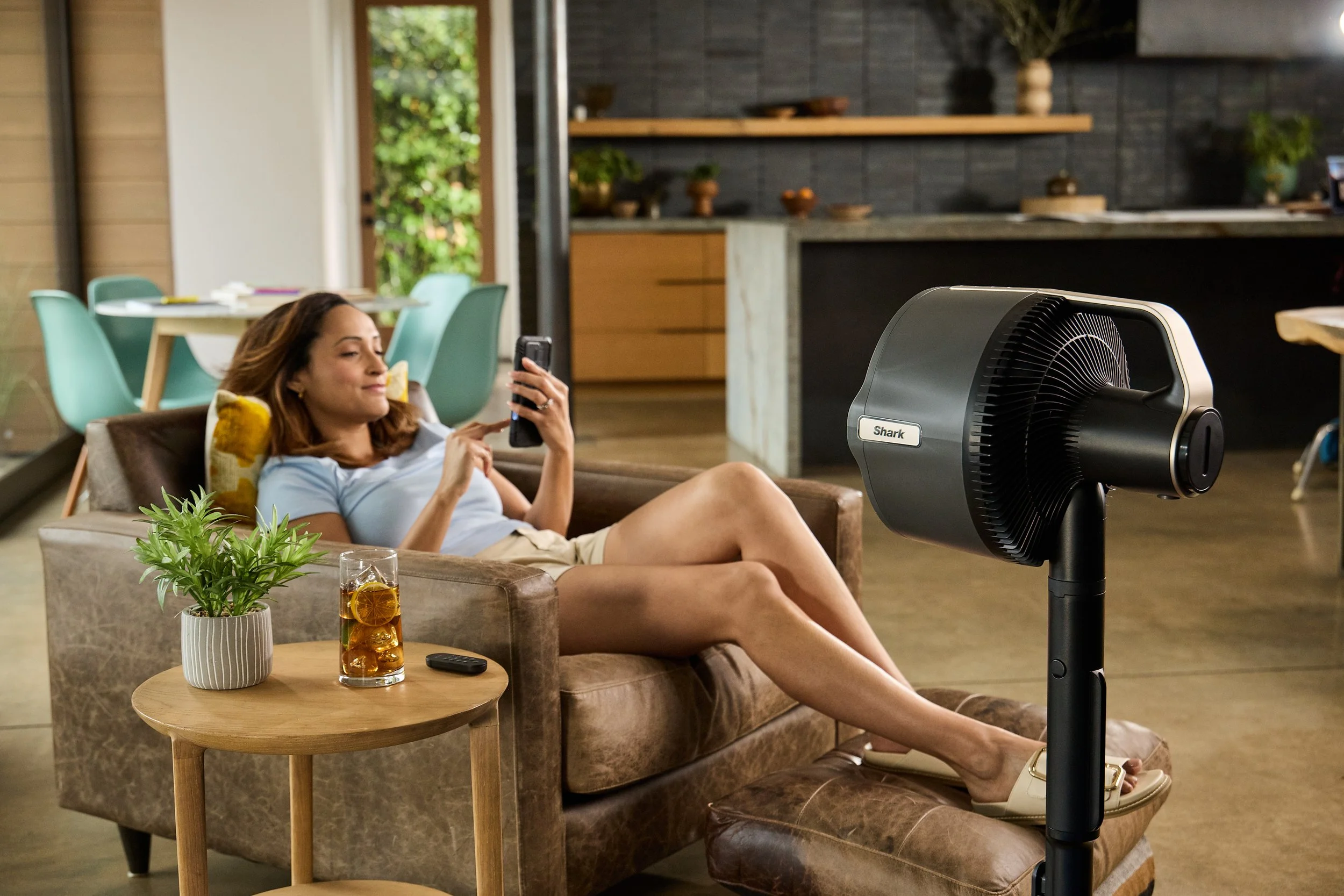 Woman relaxing with smartphone in a modern living room, sitting on a leather chair with a stand fan nearby. A small table holds a plant and a glass of iced tea with a lemon slice. The room features contemporary furniture and decor, including visible 