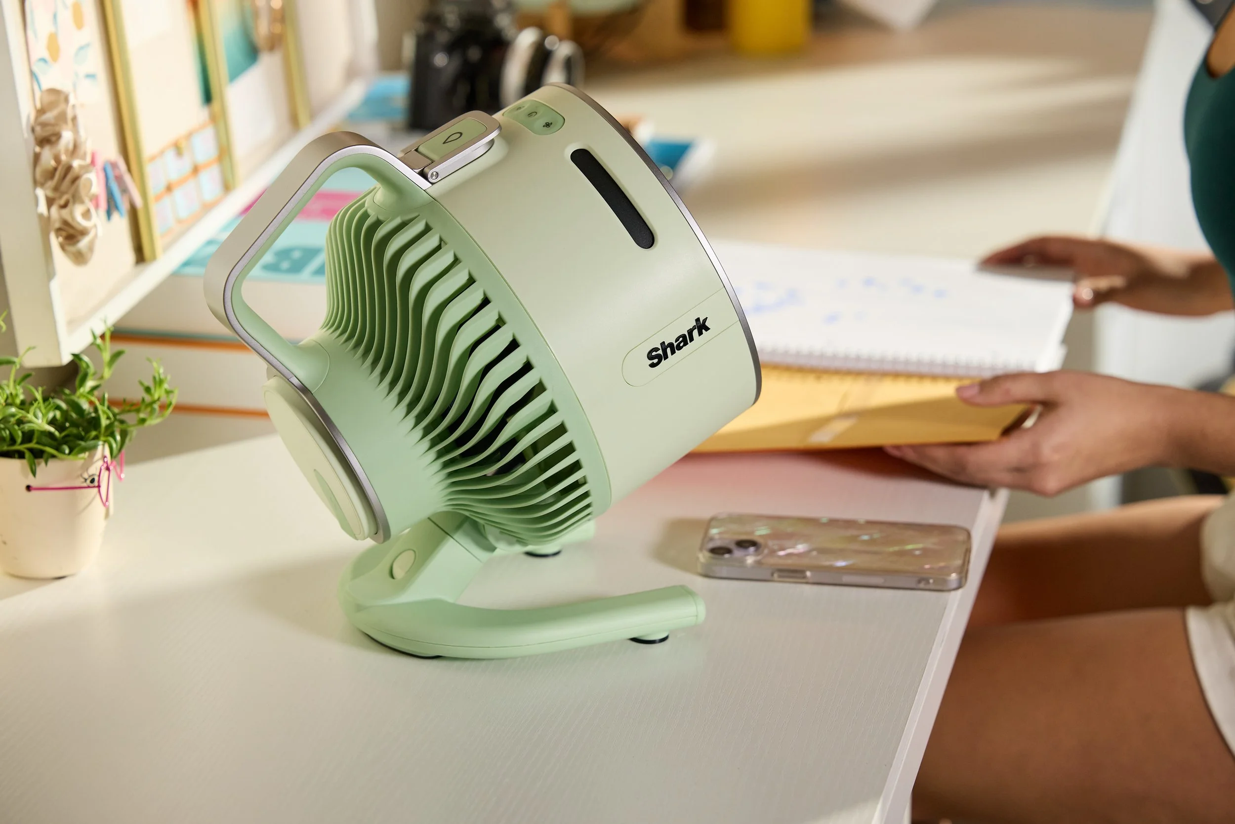 A mint green Shark brand desk fan on a white desk, next to a smartphone with a transparent case and a small potted plant. In the background, a partial view of a person holding a notebook and an envelope.