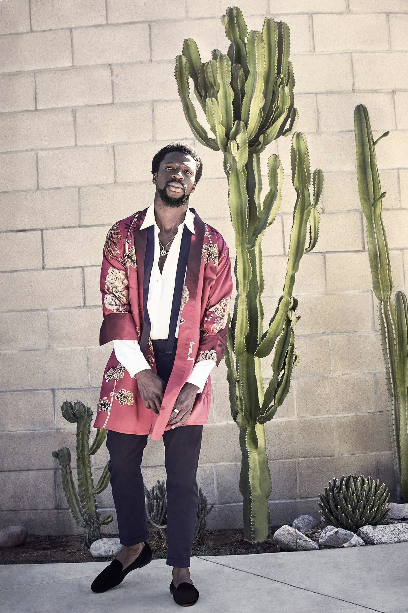 Man in floral jacket standing by cacti against brick wall.