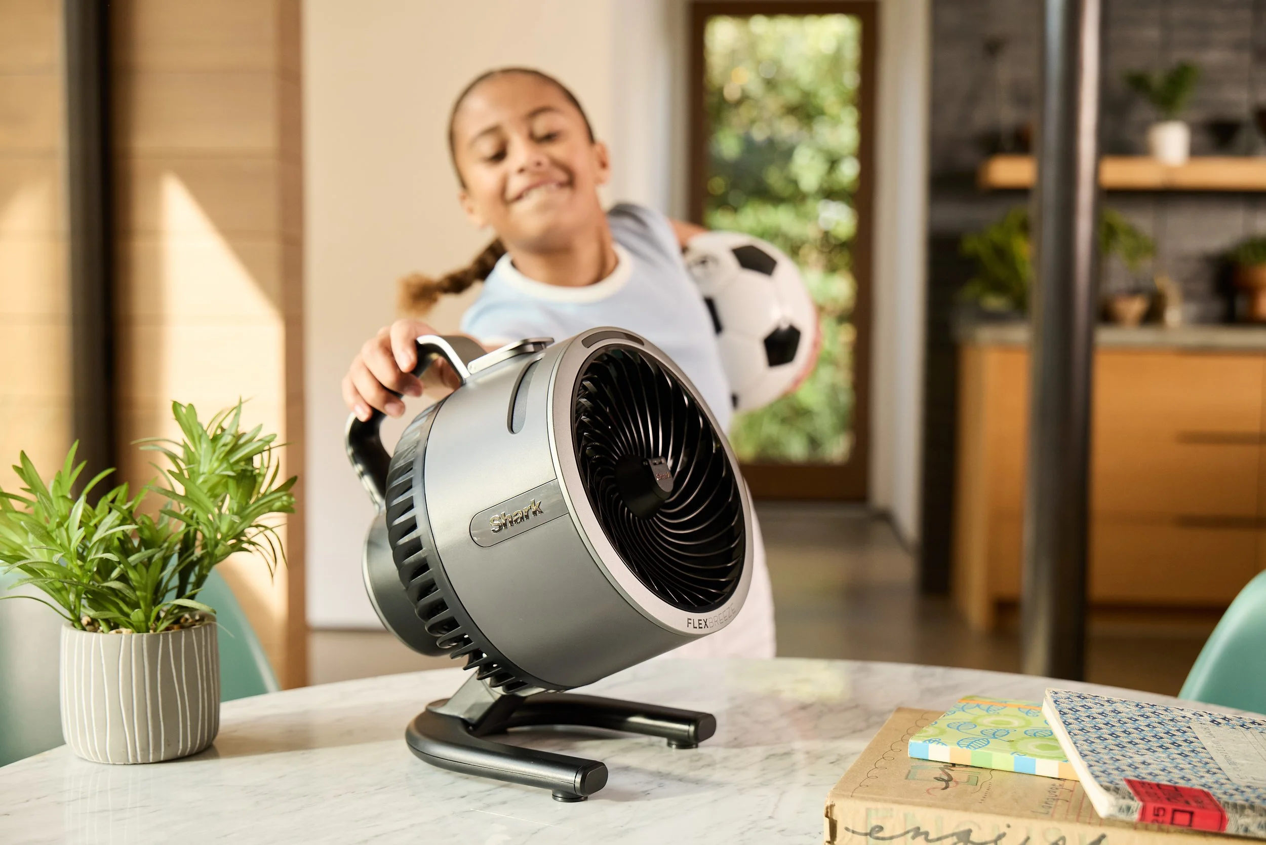 A child smiling while holding a soccer ball and turning on a Shark brand fan placed on a table with some potted plants and notebooks in a bright room.