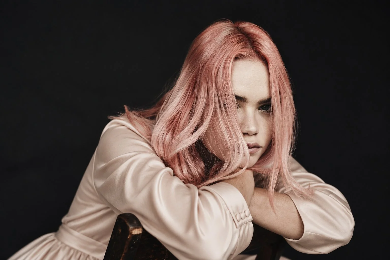 Portrait of a woman with pink hair in a pale satin jacket, leaning on folded arms against a black background.