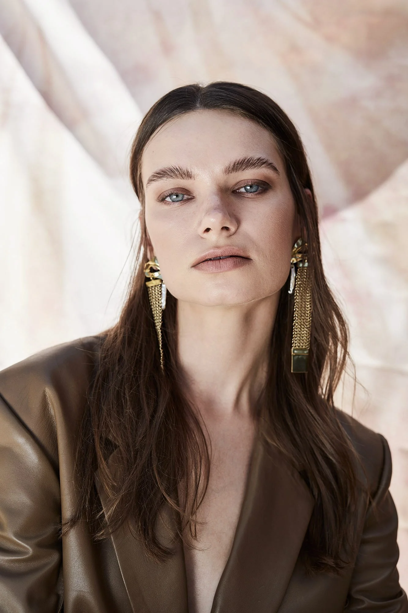 Close portrait with long gold earrings and a brown jacket against a pink backdrop