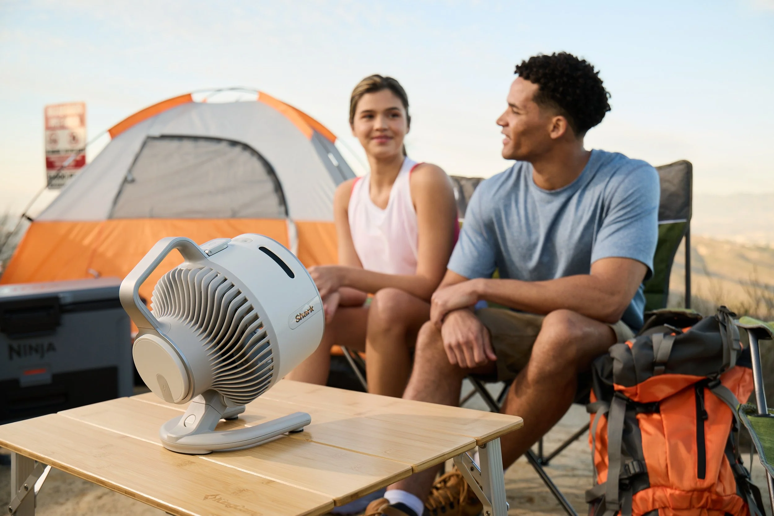 Two people sitting near a tent with camping gear and a portable fan on a table.