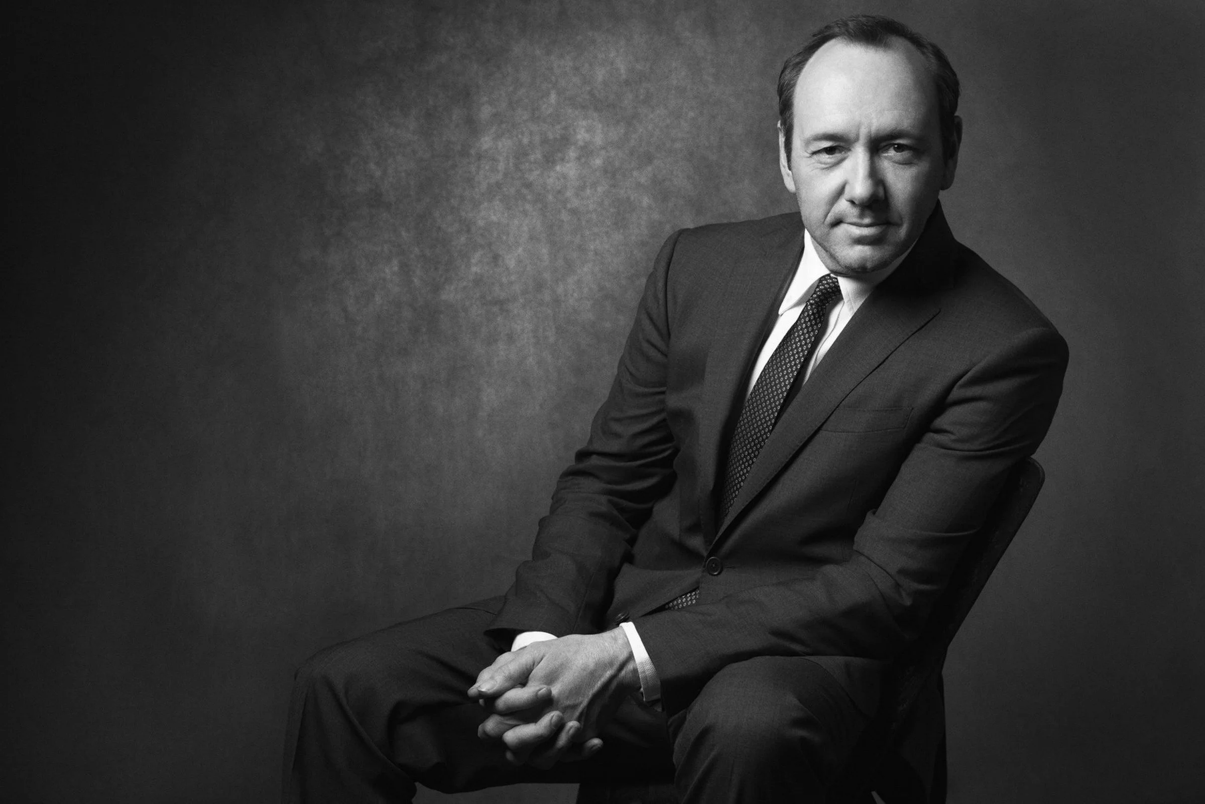 Black-and-white portrait of a seated man in a dark suit and patterned tie, hands clasped in his lap against a textured backdrop.