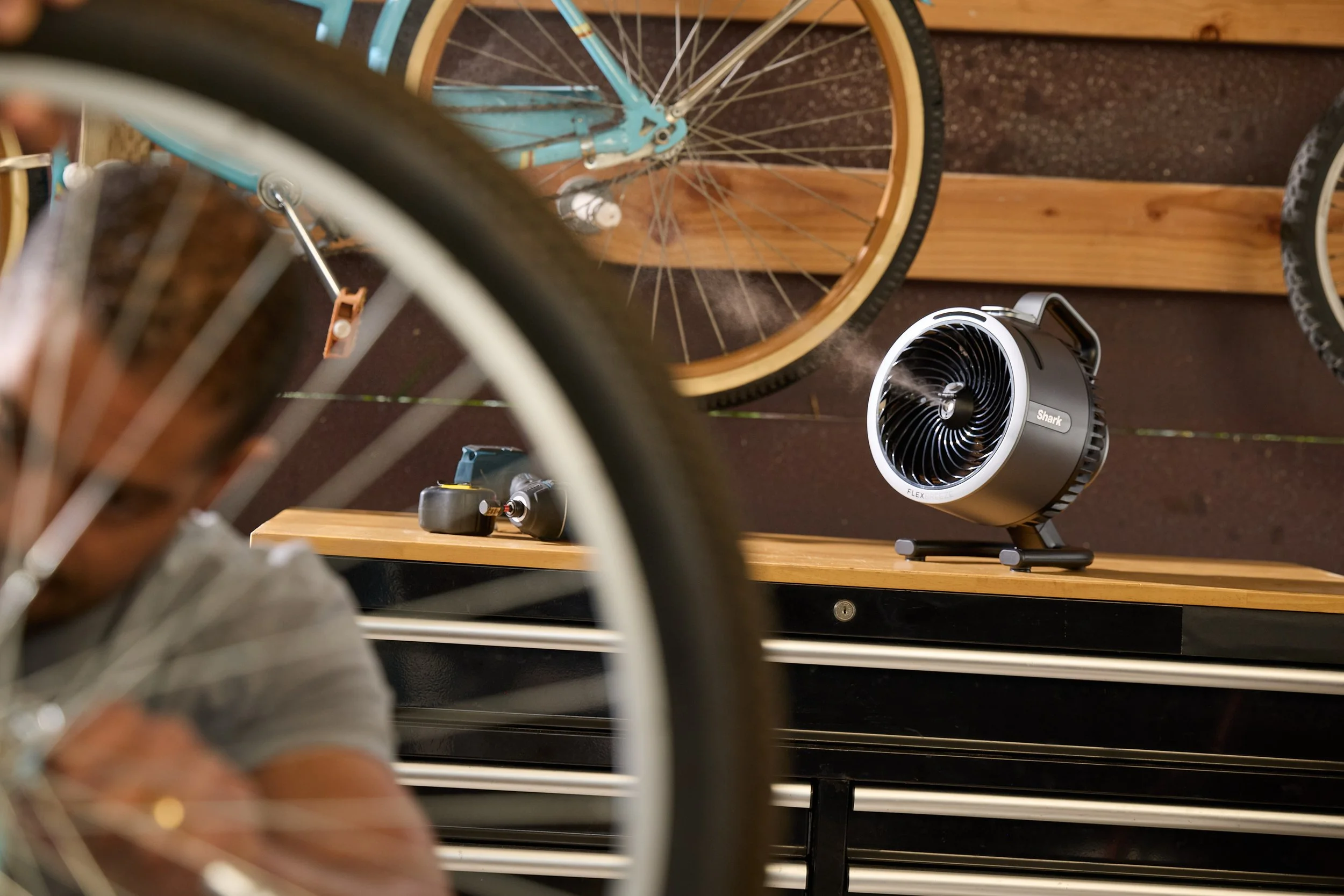 Person working on a bicycle tire in a workshop, with a fan and tools on a workbench. Bicycles are mounted on the wall.