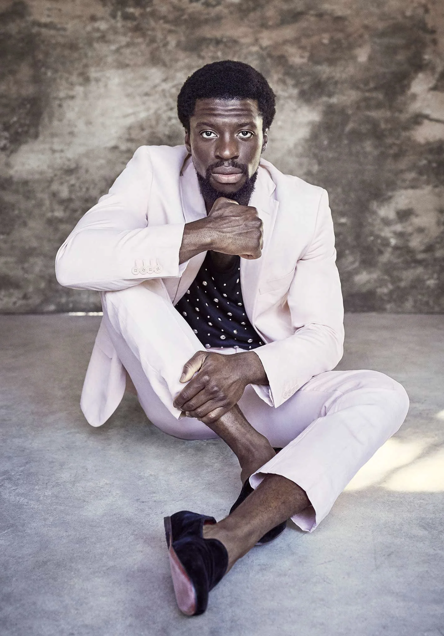 Man in light suit sitting on the floor, looking at the camera with a thoughtful expression. Background is a textured wall.