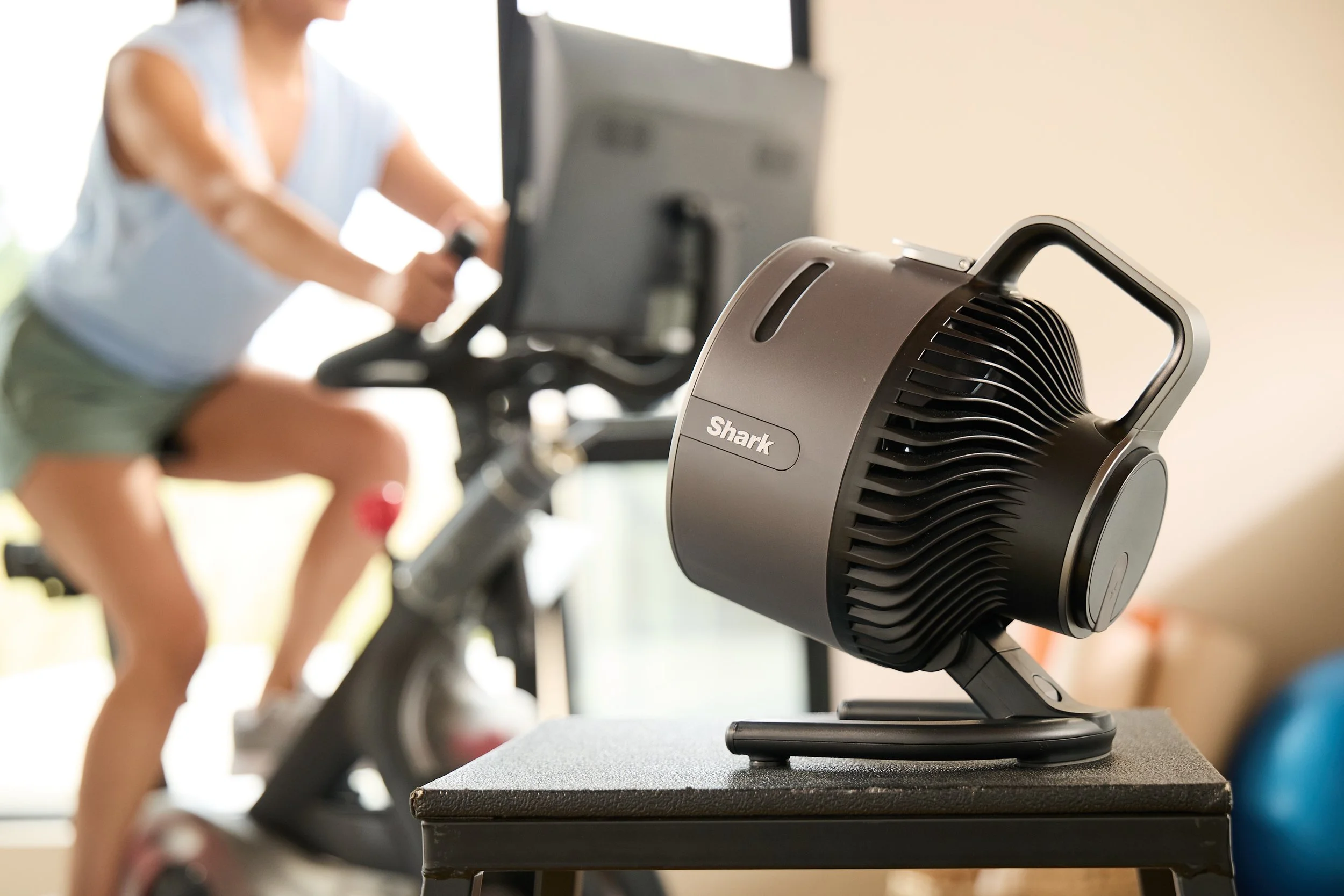 Close-up of a Shark brand fan on a table with a person exercising on a stationary bike in the background.