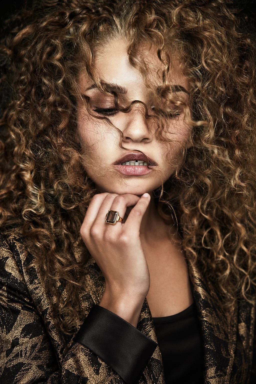 Close portrait with curls, a gold-patterned jacket and a hand beneath the chin