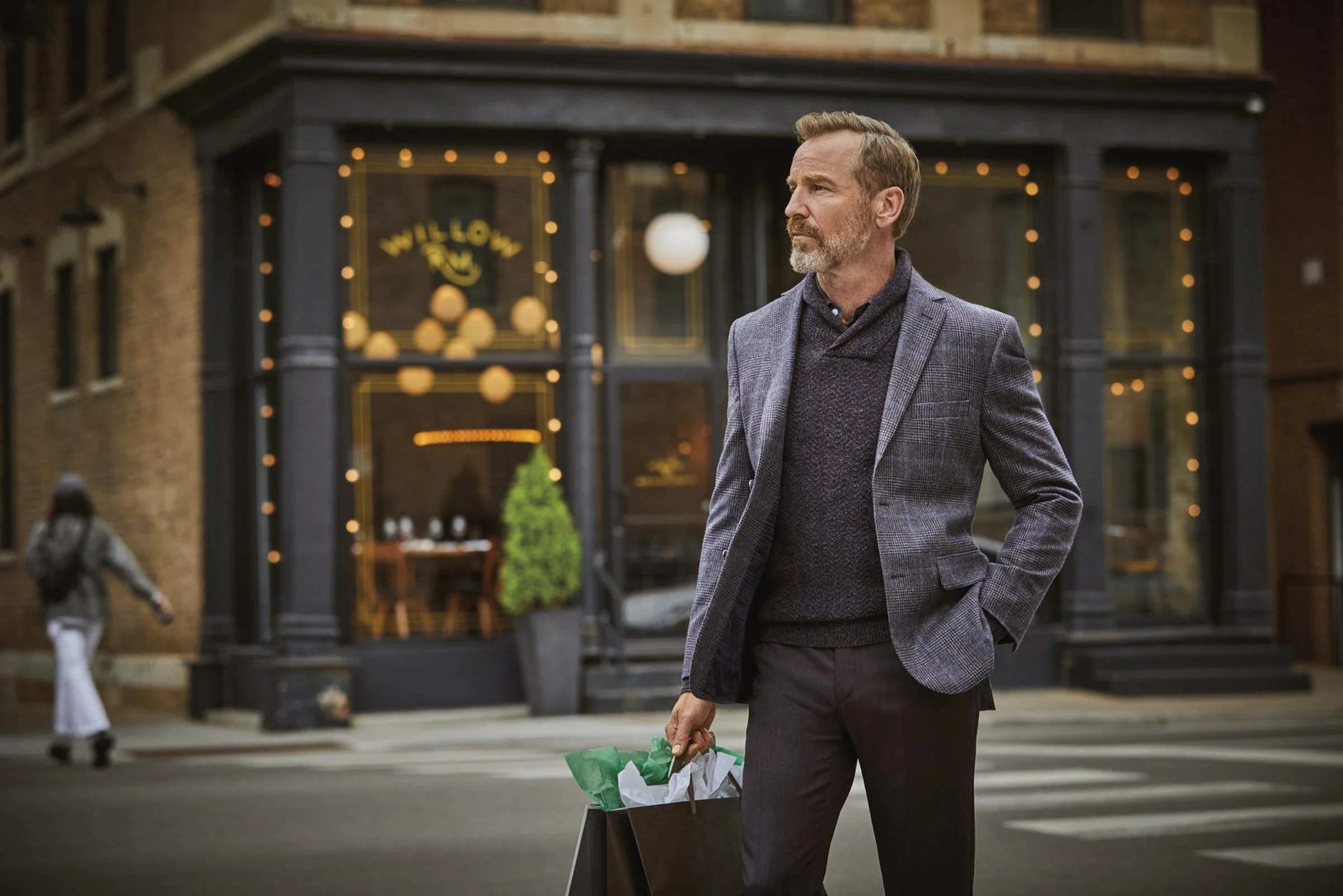 A bearded man wearing a textured blazer and dark sweater stands on a city street holding a shopping bag, with a warmly lit storefront behind him.