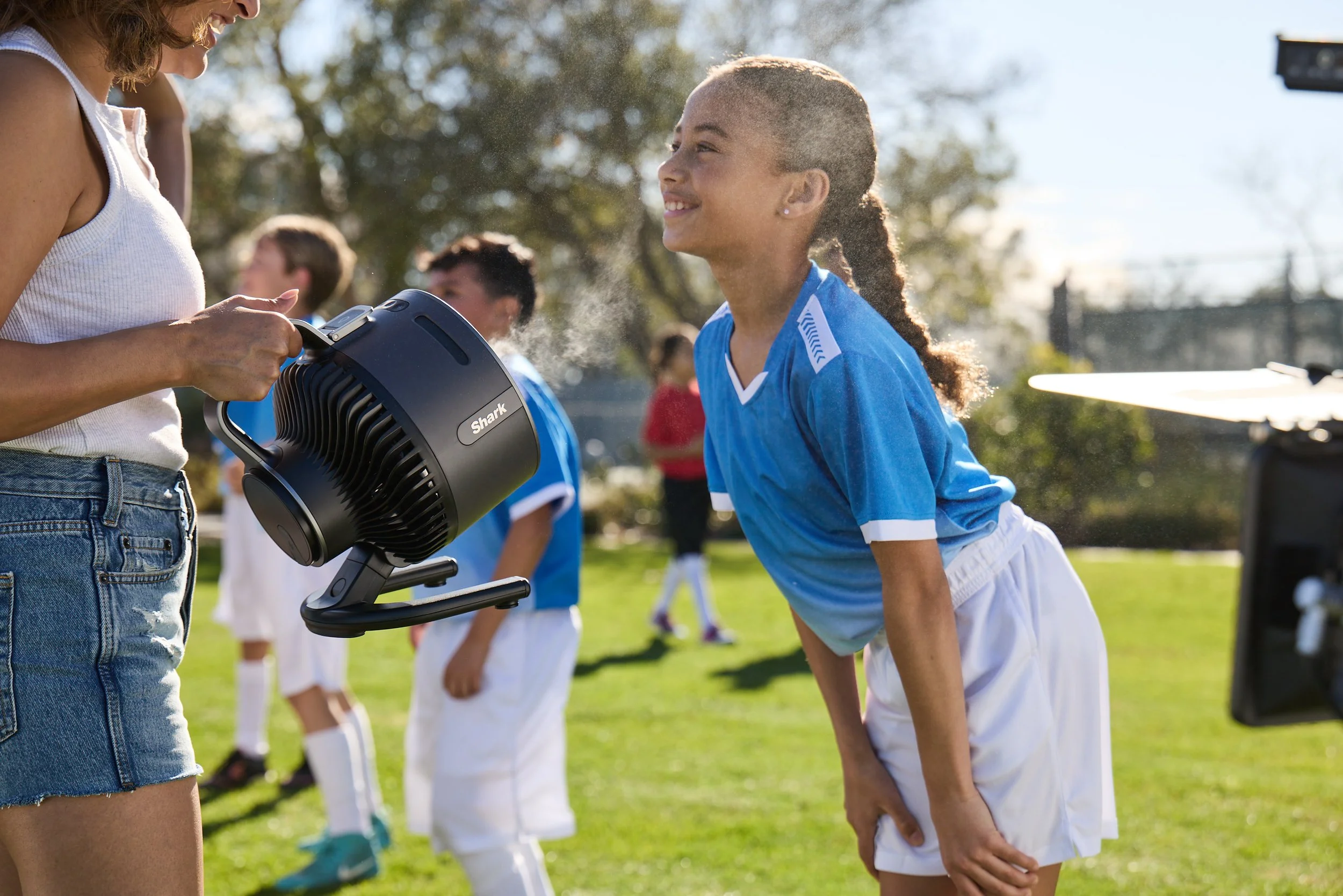 A person using a Shark handheld mister to spray water on a smiling girl wearing a blue sports jersey on a sunny soccer field.