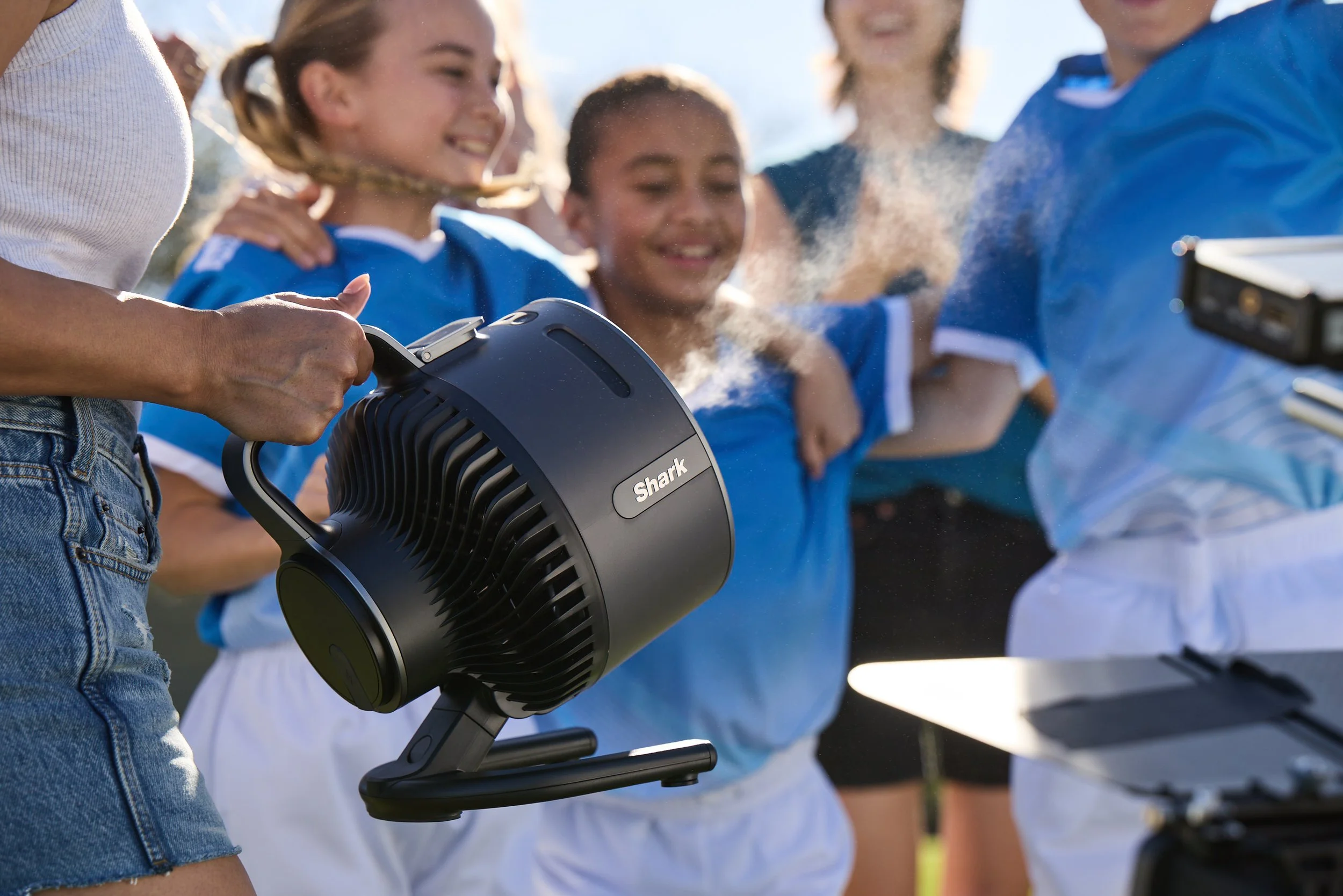 Person holding a Shark handheld device spraying mist near children in blue sports jerseys outdoors.