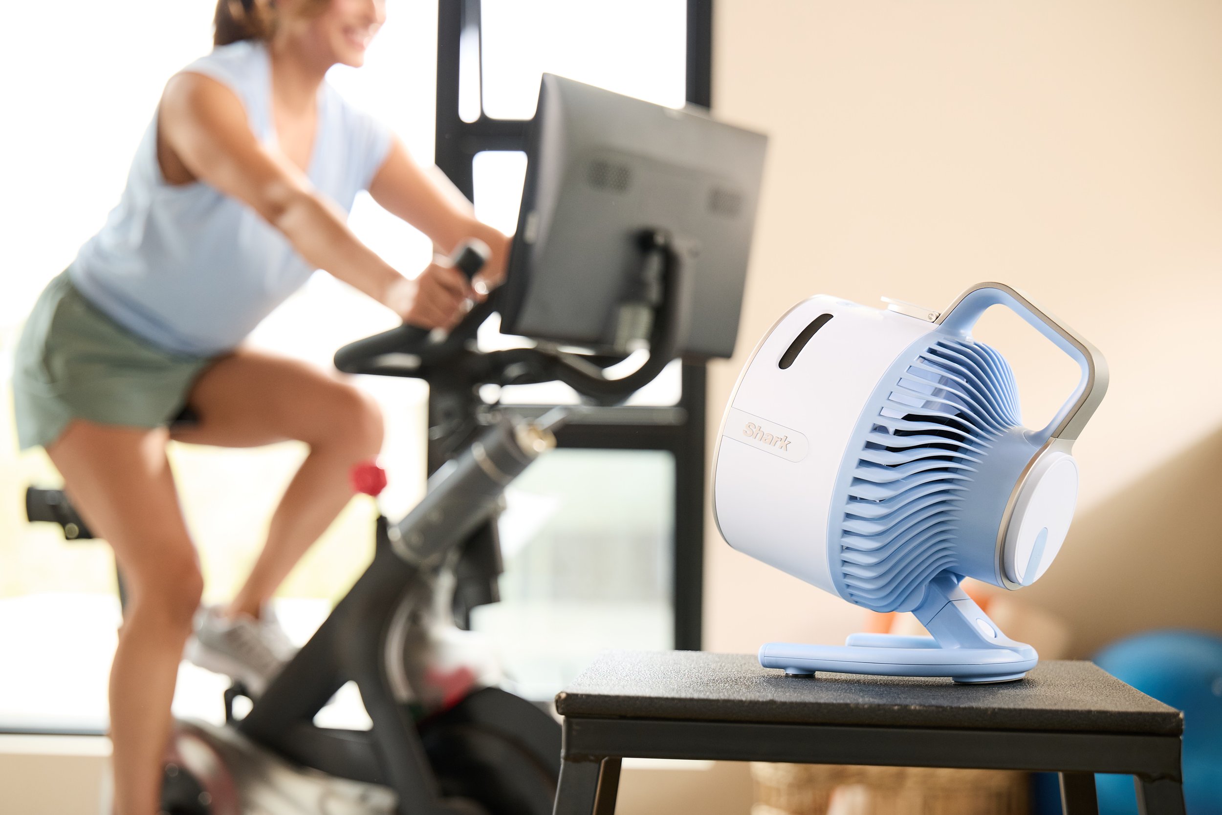 Woman exercising on a stationary bike with a fan on a table nearby.