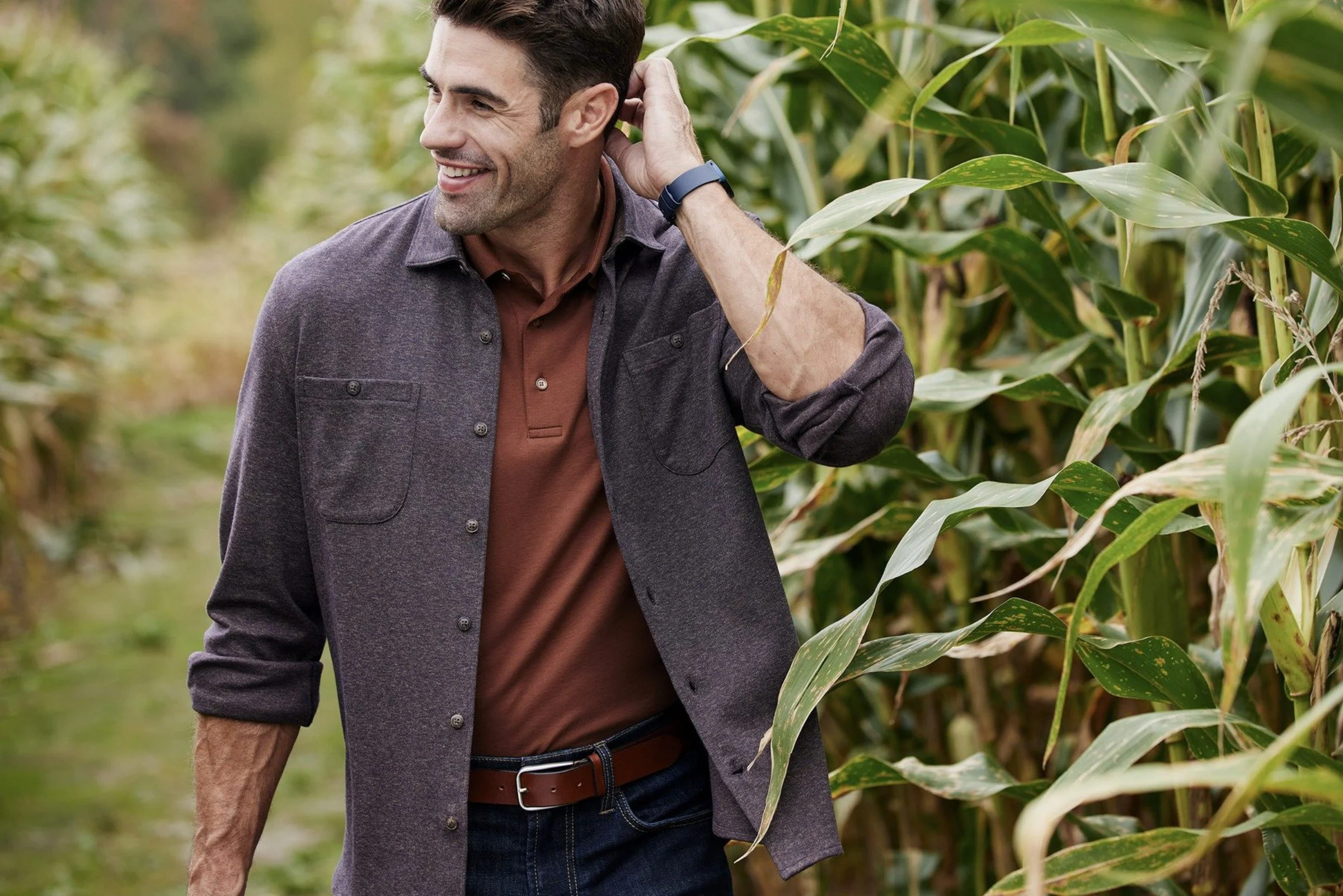 A smiling man in a charcoal overshirt and rust-colored polo stands beside tall corn plants, with one hand raised behind his head.
