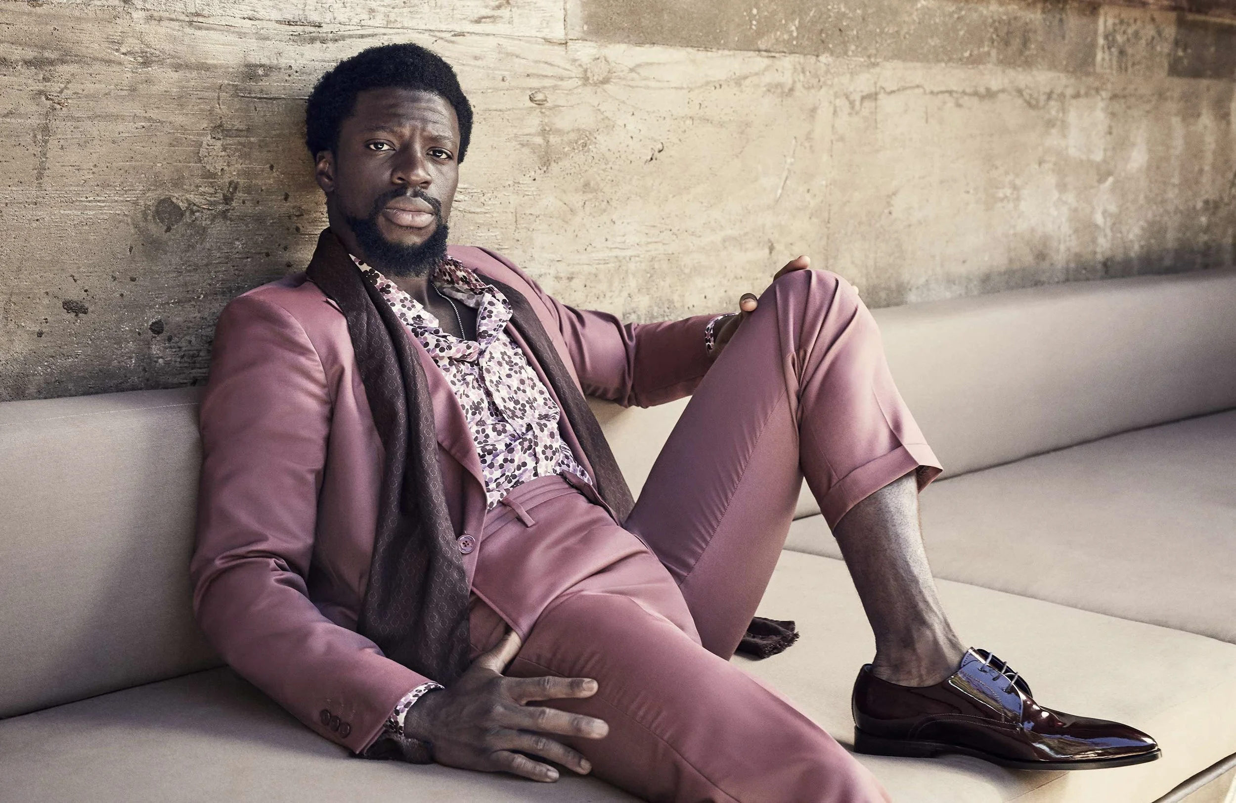 Man in stylish plum-colored suit sitting on a beige couch against a textured wall.