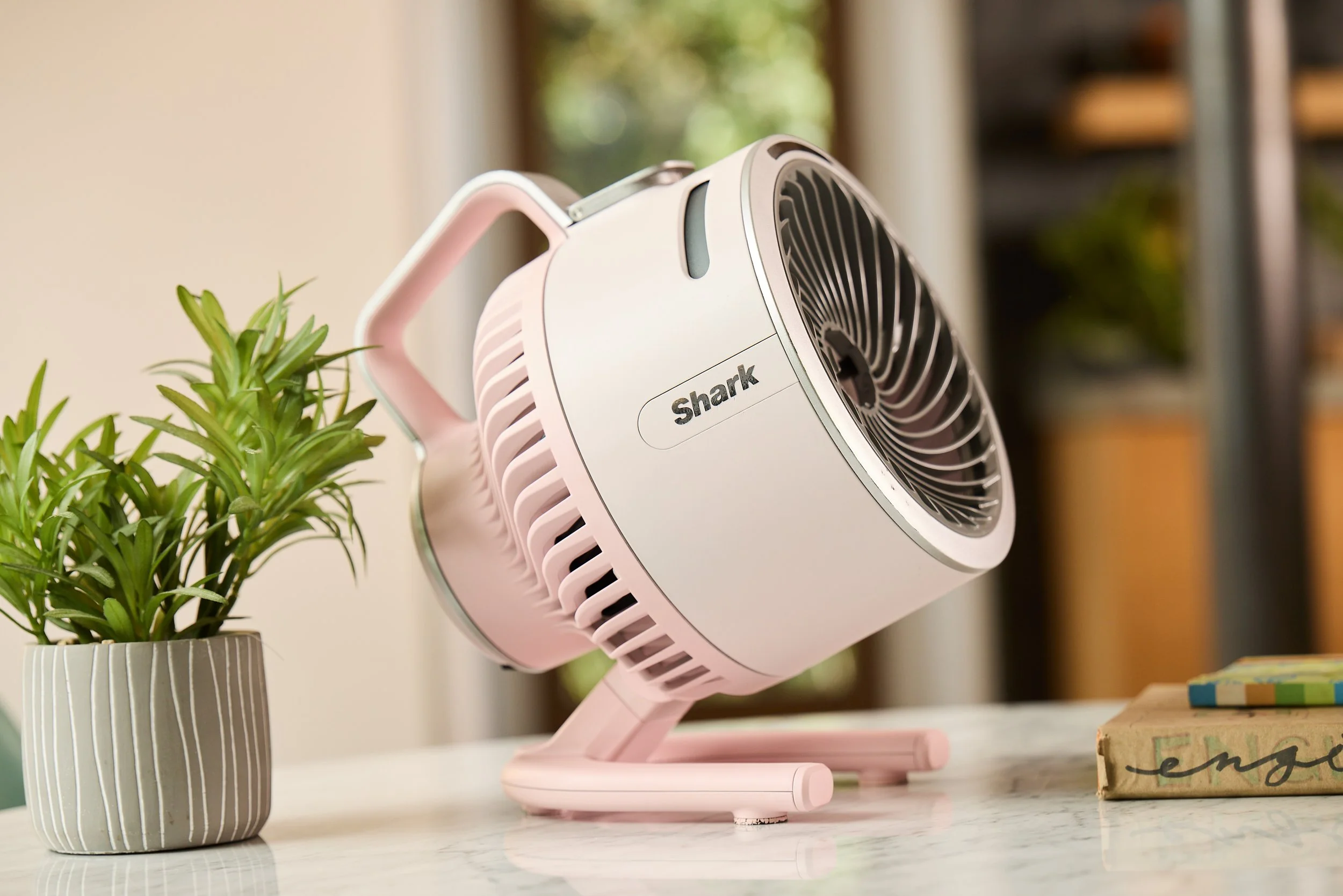 A pink Shark brand tabletop fan on a marble surface beside a small potted plant and some books.